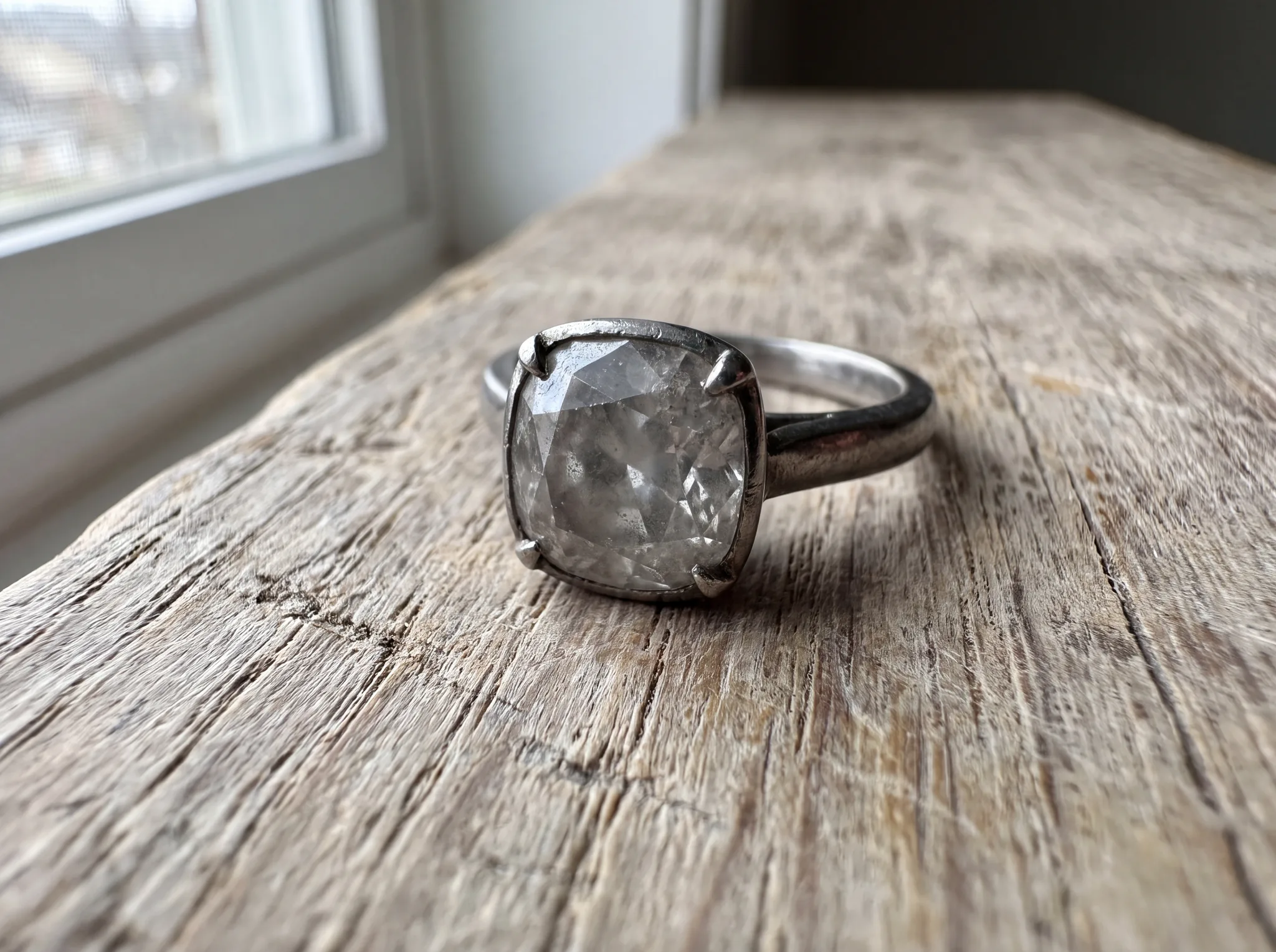 A diamond solitaire ring on pale weathered oak, the stone reading grey and flat rather than bright, its lower facets clouded with a film.