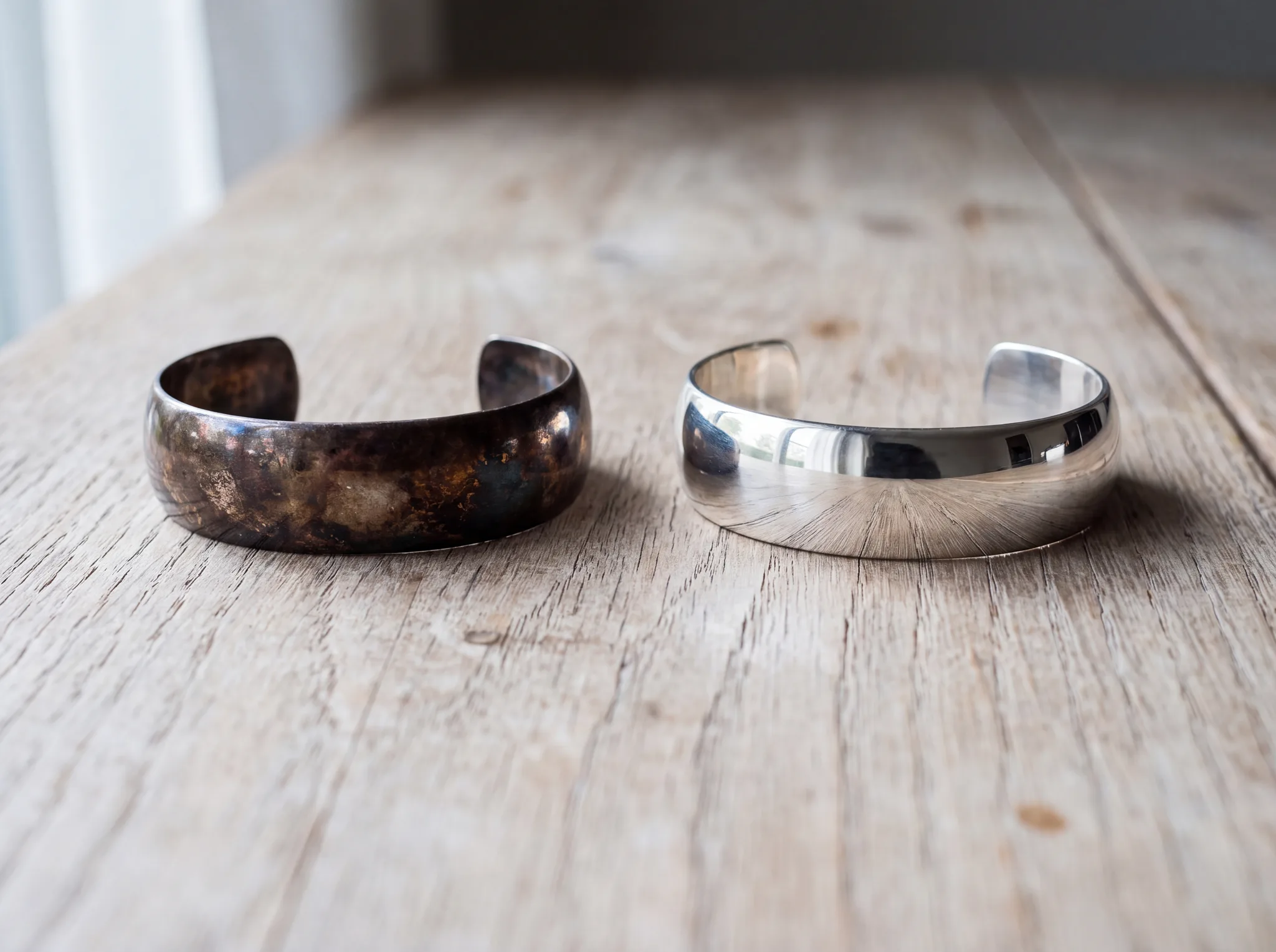Two sterling silver bangles side by side on pale weathered oak, the left one darkened brown-black with tarnish and the right one polished to a bright mirror finish.