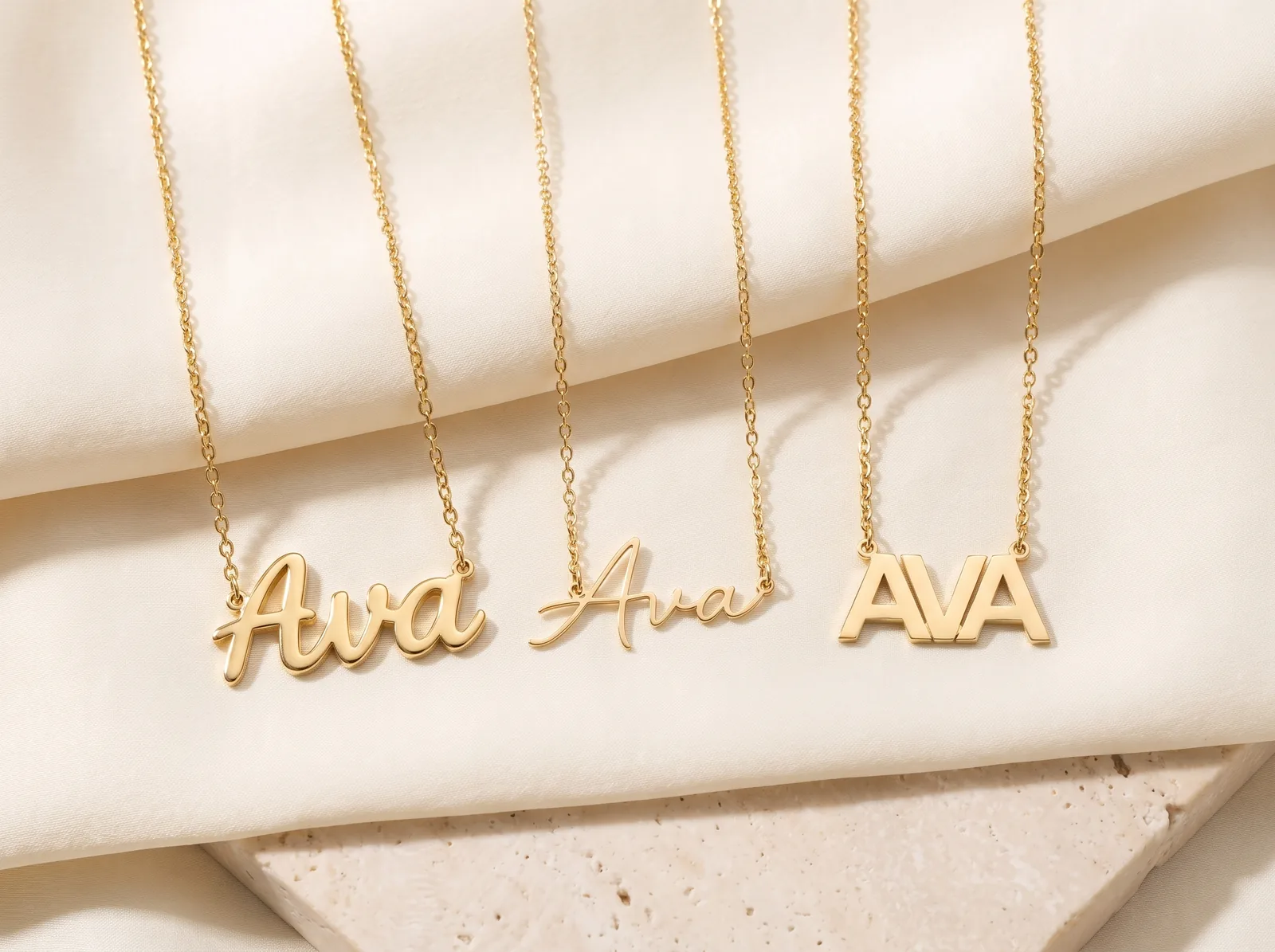 Three yellow gold name necklaces laid side by side on cream silk over pale travertine, each reading Ava: a bold joined script on the left, a much finer cursive in the middle, and separate block capitals on the right.