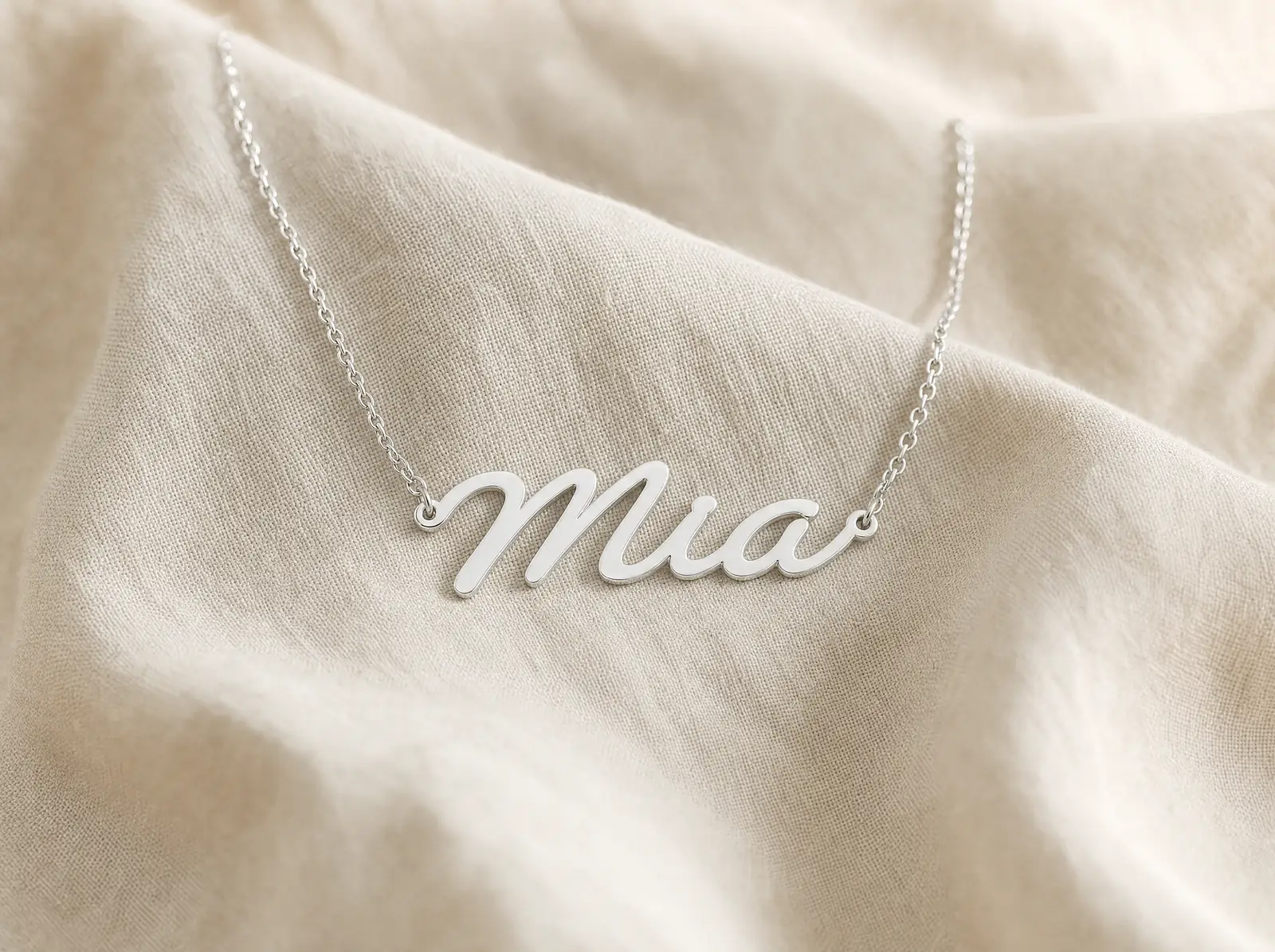 A single silver script name necklace reading Mia on a fine cable chain, laid flat on cream linen.