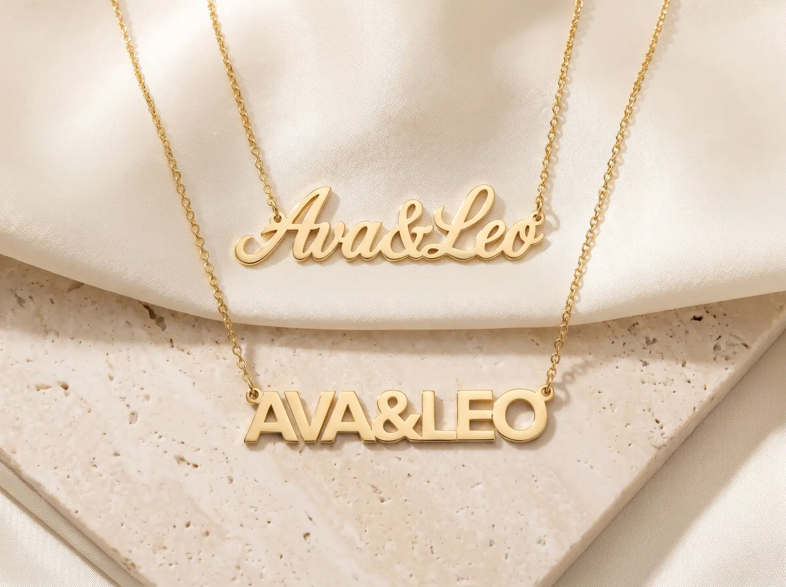 Two yellow gold two-name necklaces on cream silk and pale travertine, both reading Ava and Leo joined by an ampersand: a flowing joined script piece above, and a piece in separate block capitals below.