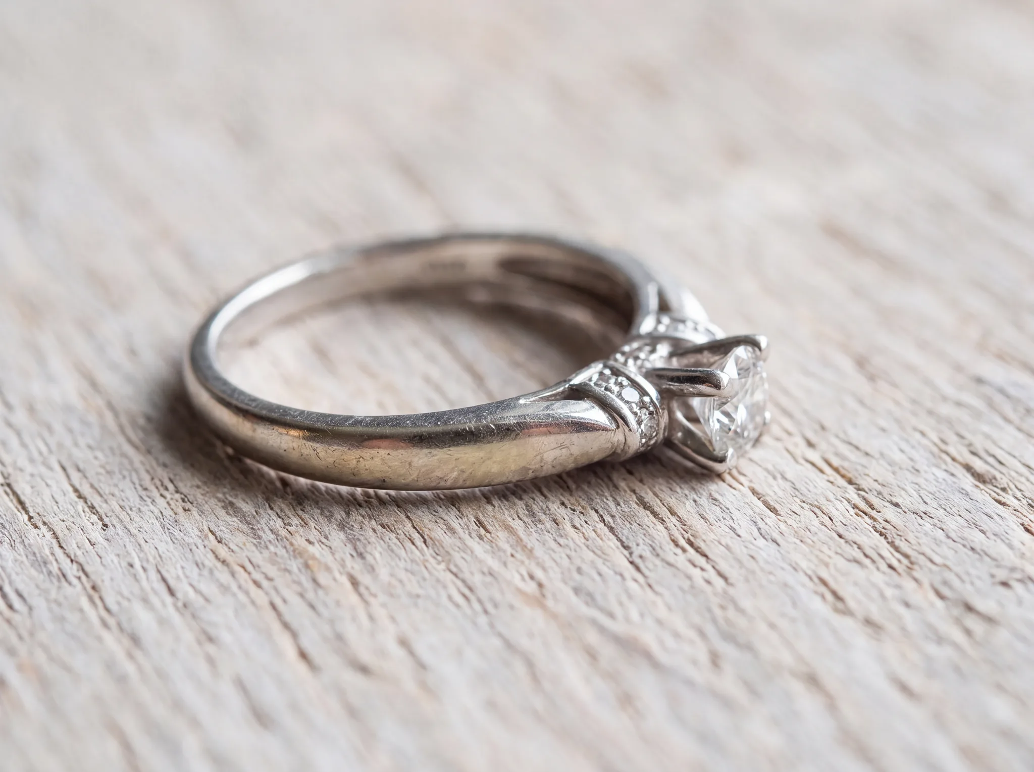 A white gold diamond ring on a pale wood surface, its shank and shoulders gone warm and faintly yellow-grey where the rhodium plating has worn through while the setting stays bright white