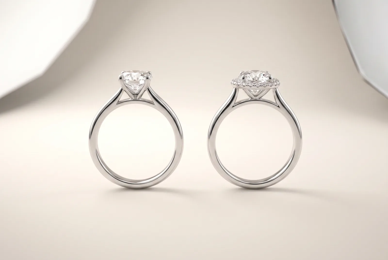 Two platinum engagement rings standing side by side, each holding an identical round diamond. The ring on the left is a plain solitaire; the one on the right has the same stone encircled by a halo of small diamonds.