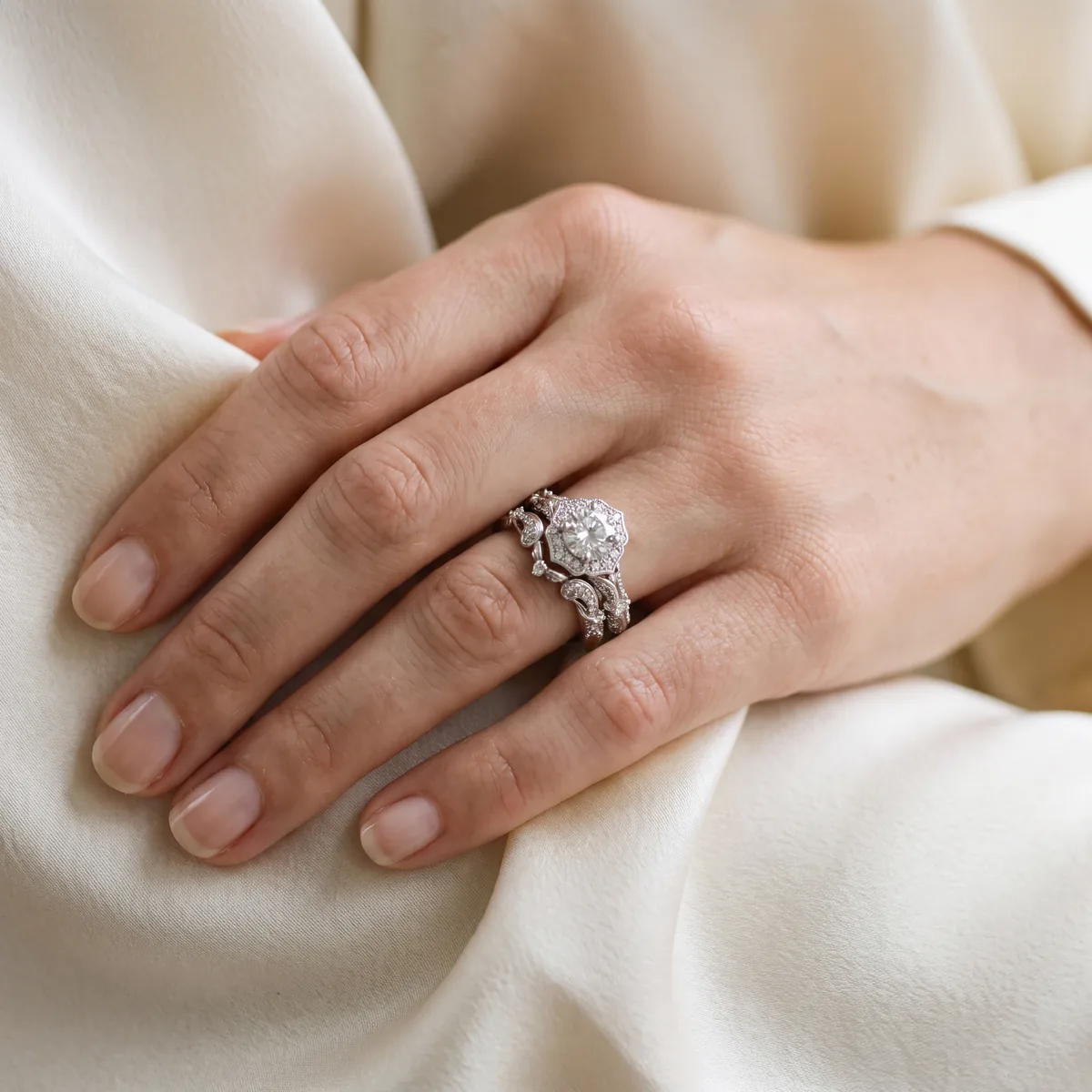 A hand resting on cream fabric, wearing a round diamond engagement ring with a halo. A curved diamond wedding band follows the shape of the halo and sits close against it.