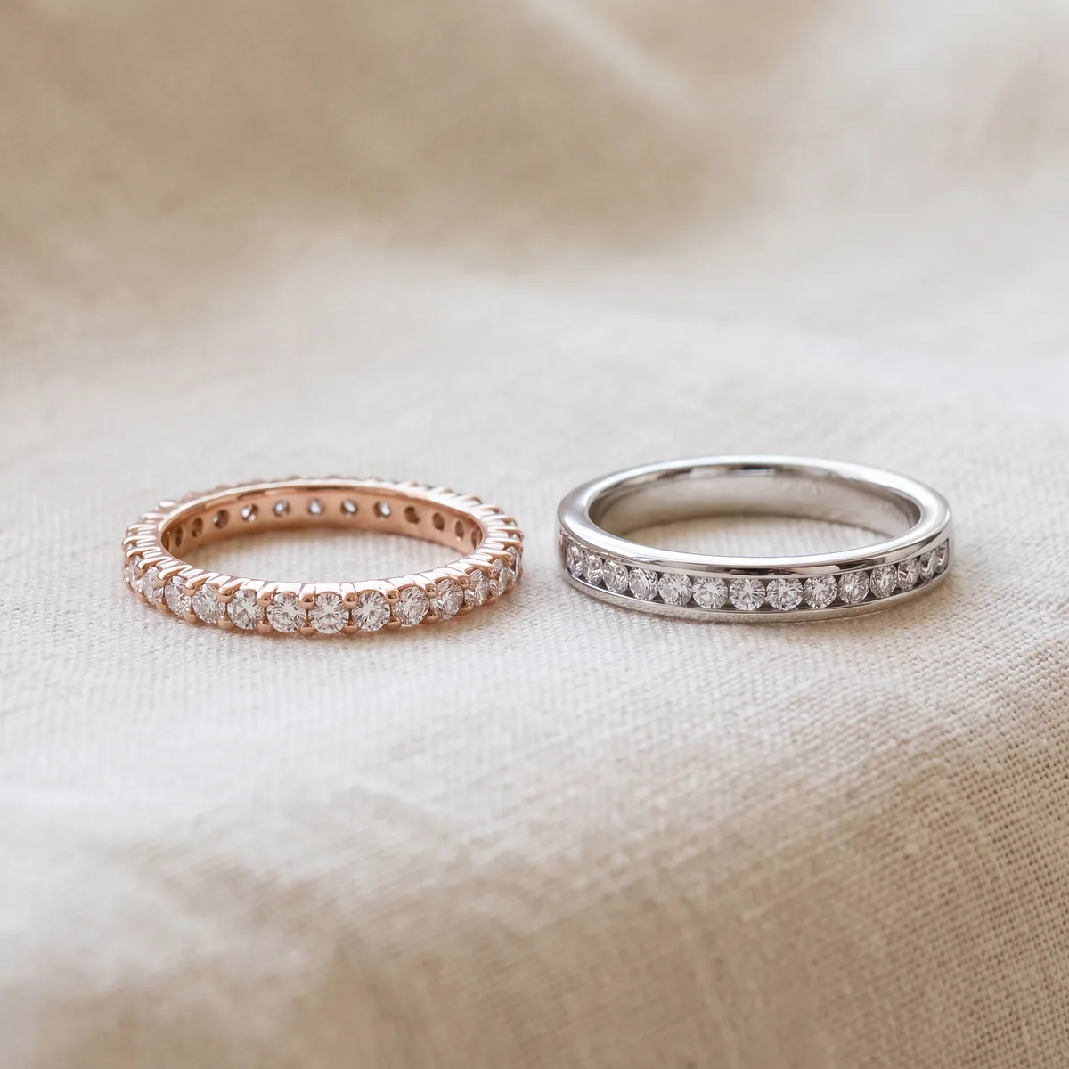 Two diamond bands standing side by side on cream linen. On the left, a rose gold band with round diamonds all the way around. On the right, a white metal band with round diamonds set in a channel across the top.