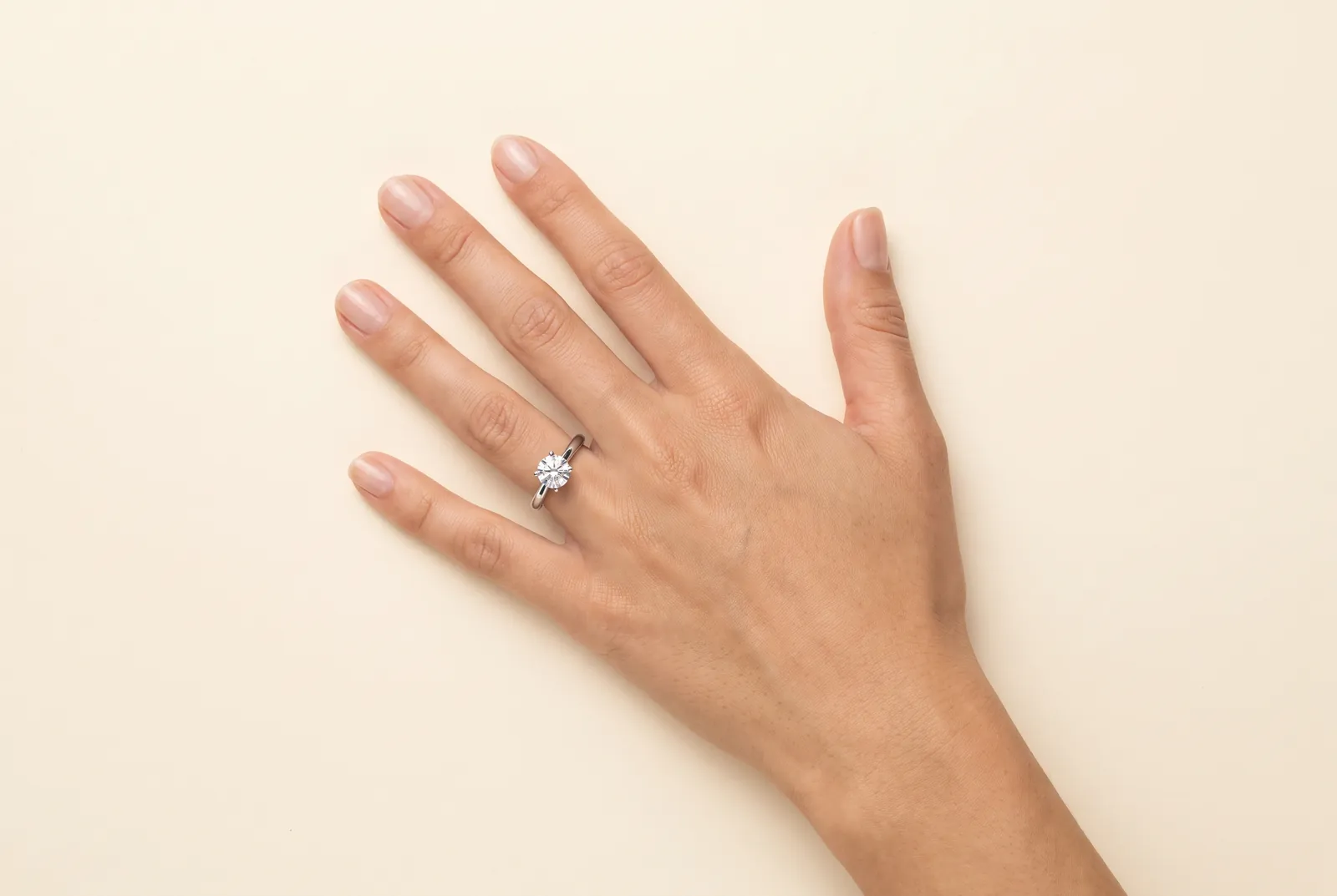 A left hand resting palm-down on a cream backdrop with the fingers and thumb spread, wearing a platinum solitaire with a single round diamond on the fourth finger.