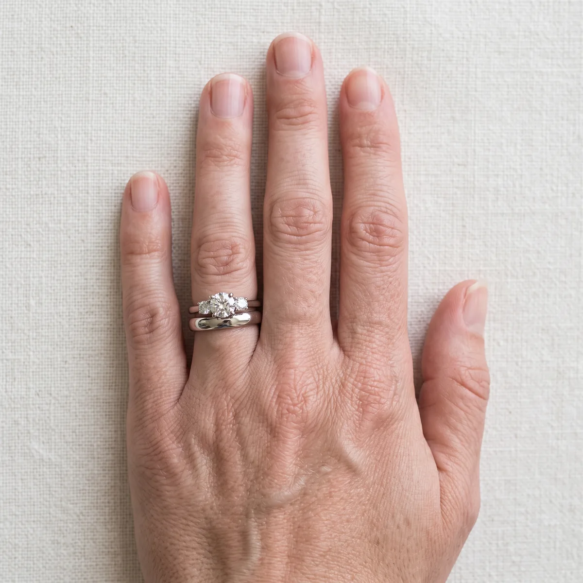 A left hand with a plain white gold wedding band at the base of the ring finger and a three-stone engagement ring above it, toward the fingertip.