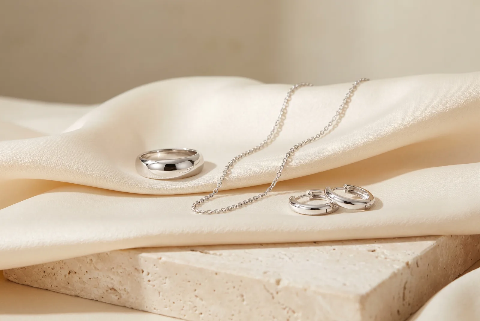 A domed white gold band, a fine white gold cable chain and a pair of white gold huggie hoop earrings on cream silk over a pale stone slab.