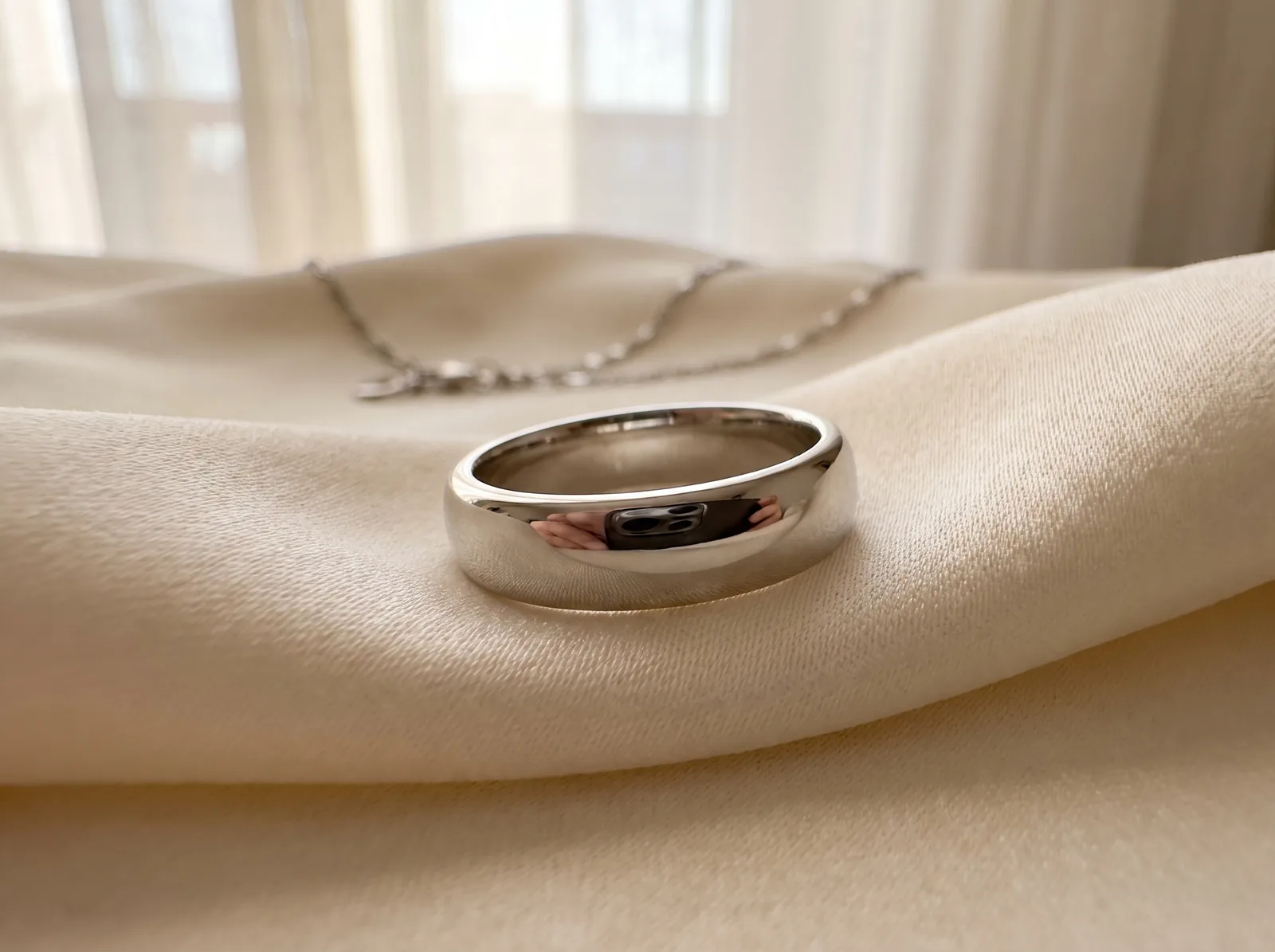 A wide, mirror-polished white gold band beside a sculptural white gold pendant on a fine chain, on a pale travertine slab in window light.
