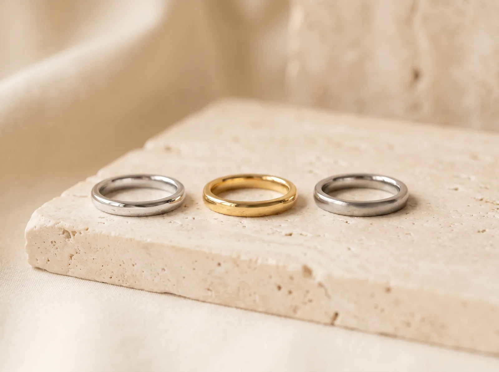 Three plain domed bands side by side on a pale stone slab: white gold on the left, yellow gold in the middle, platinum on the right.