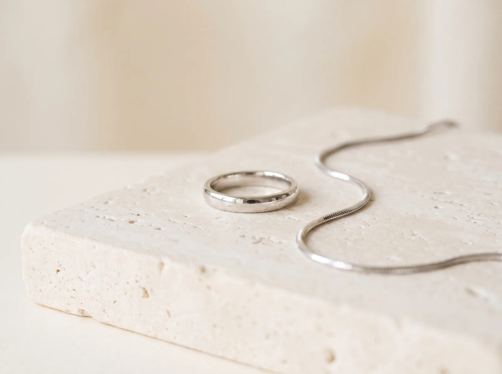 A polished white gold band and a fine white gold snake chain on a travertine slab in window light.