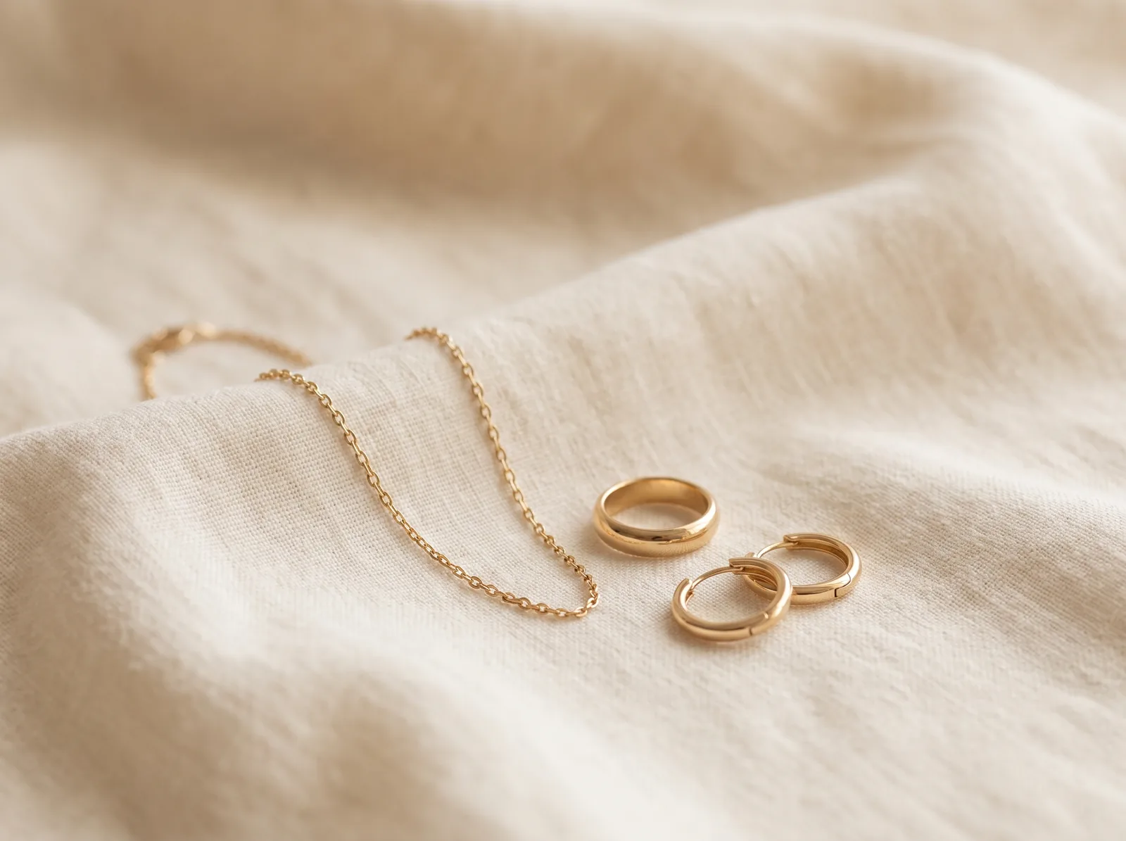 A fine yellow gold cable chain, a domed yellow gold band and a pair of yellow gold huggie hoop earrings on cream linen.