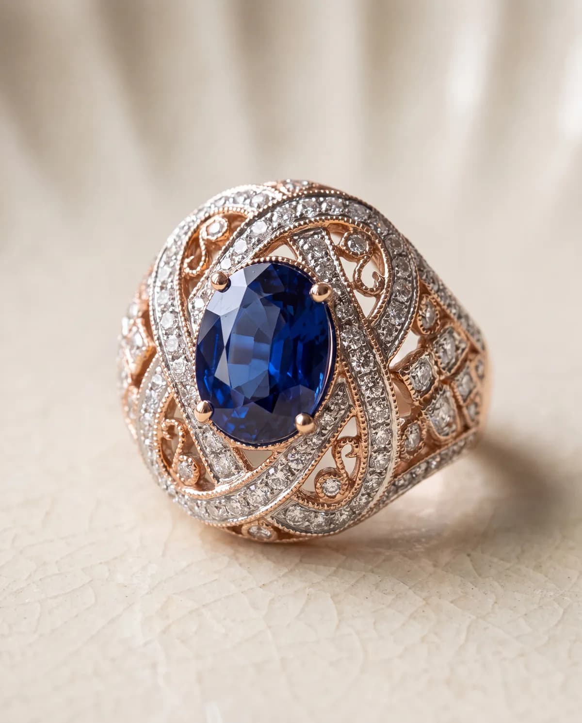 A rose and white gold filigree ring set with an oval blue sapphire surrounded by pavé diamonds, on a cream textured surface