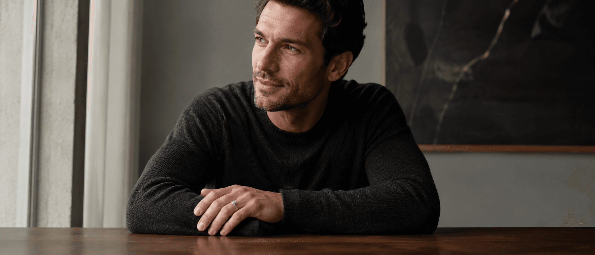 Man in a charcoal sweater at a walnut table wearing a brushed platinum bezel-set diamond engagement ring