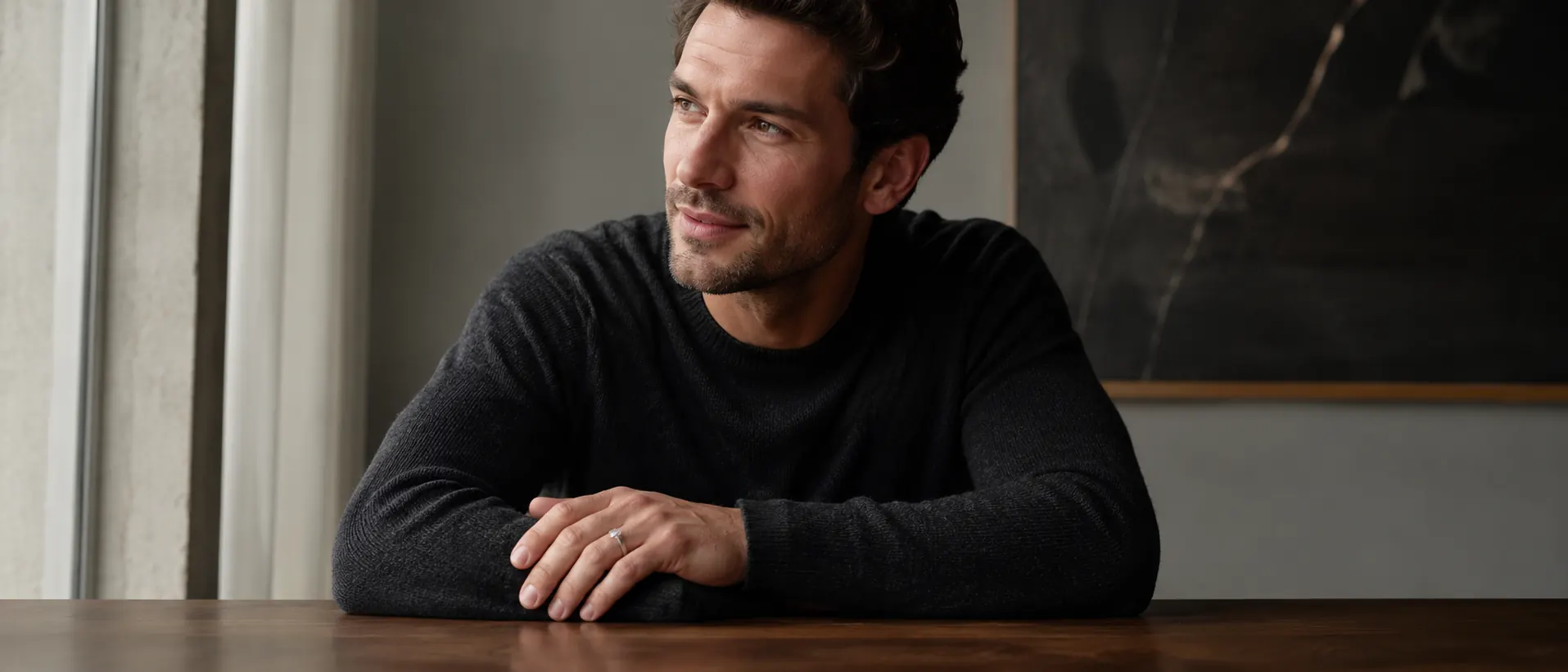 Man in a charcoal sweater at a walnut table wearing a brushed platinum bezel-set diamond engagement ring