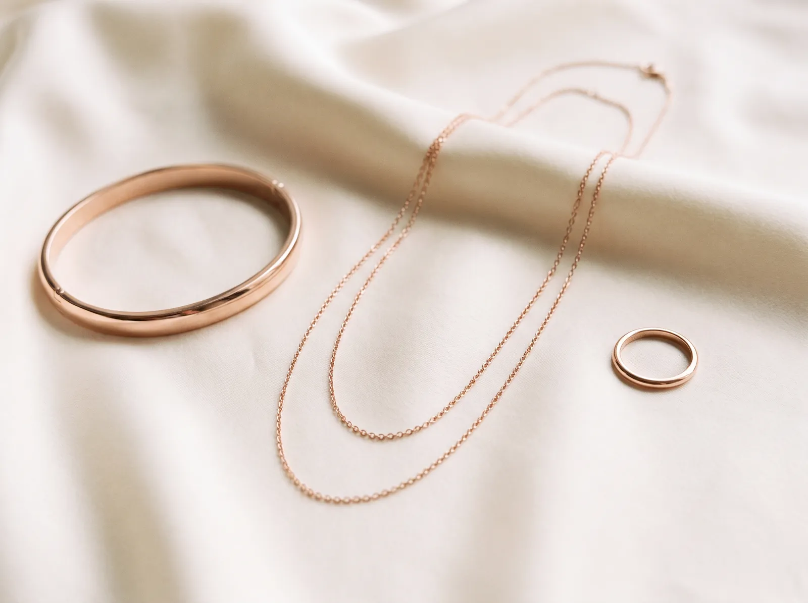 A rose gold bangle, two layered rose gold chains and a rose gold stacking ring on cream silk