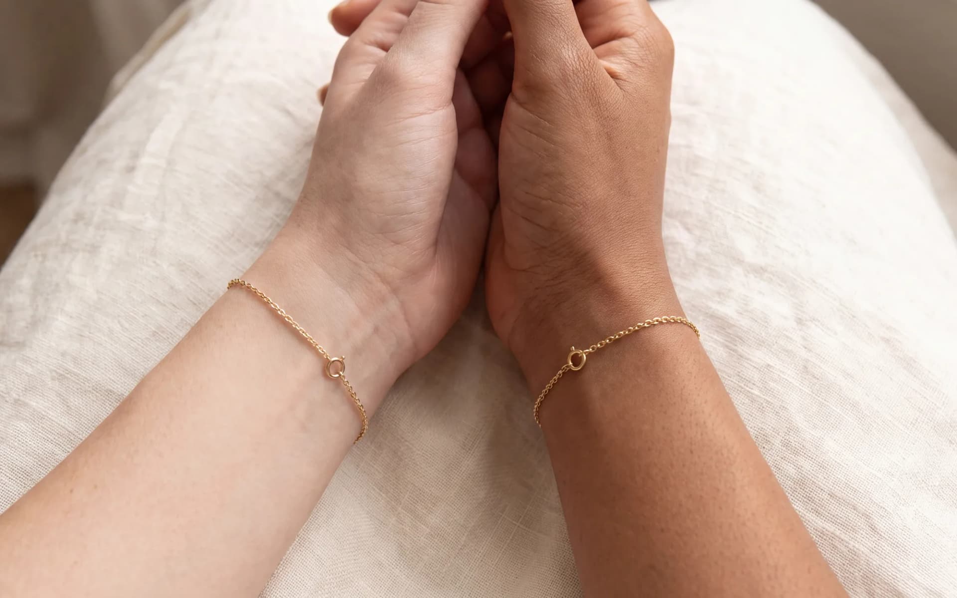 Two wrists in different skin tones wearing matching fine chain bracelets, fingers loosely linked