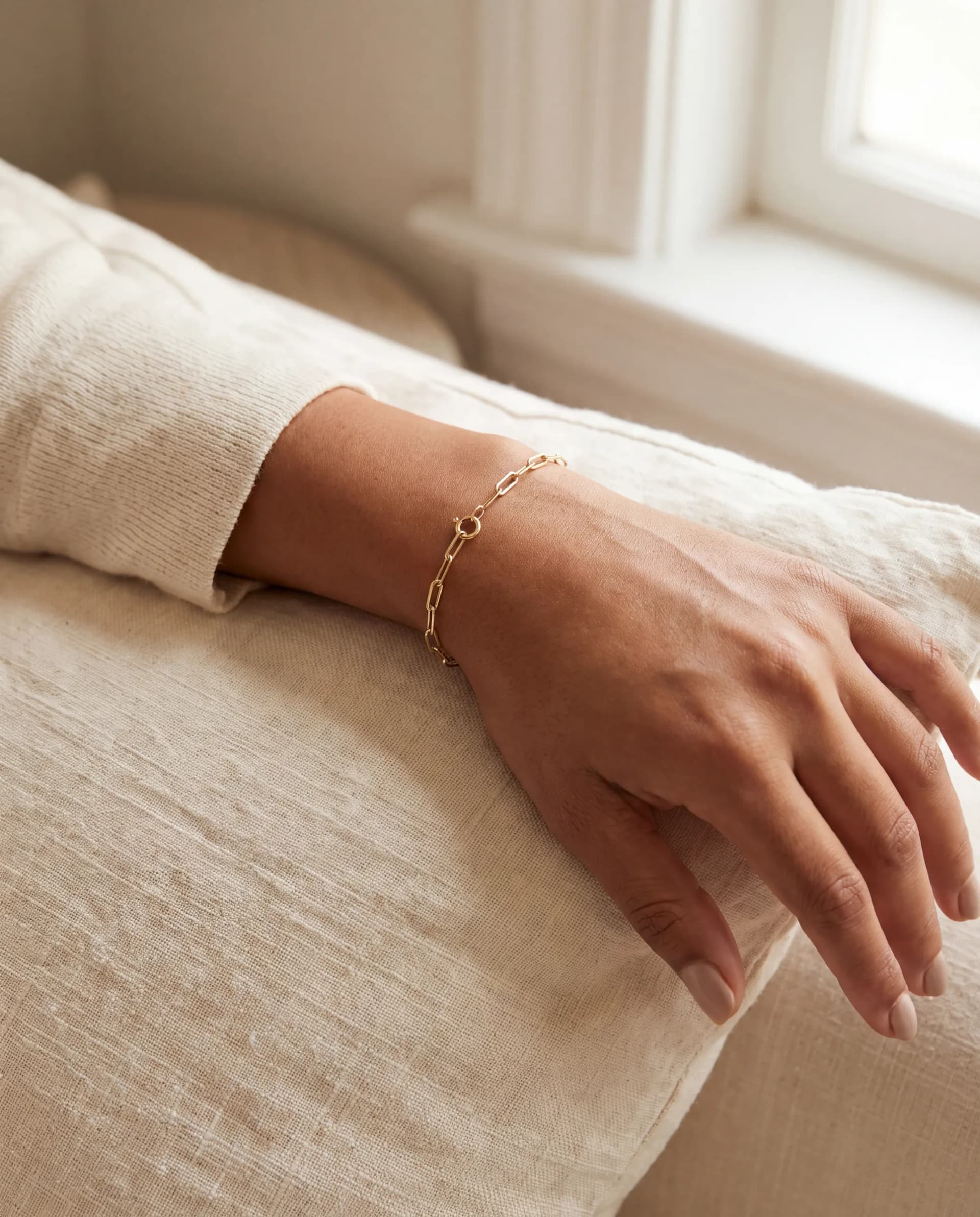 A single wrist wearing a fine 14K gold paperclip chain bracelet with the sleeve pushed back, on ivory linen