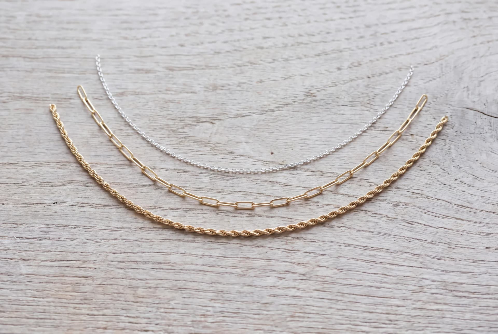 Three fine chains of different thickness laid side by side on an oak surface, one silver-colored and two gold-colored