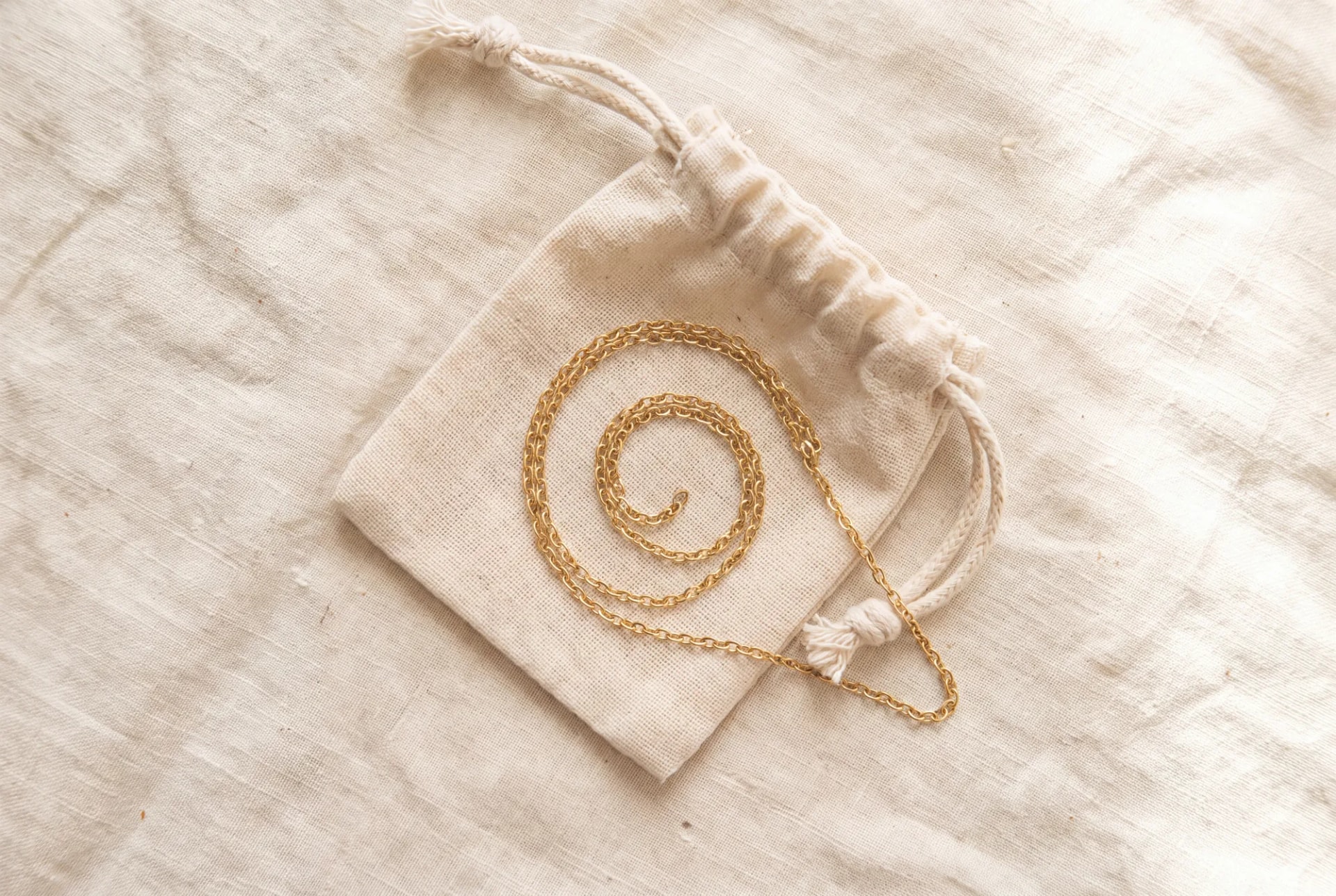 A fine chain bracelet lying open on linen beside a small folded cotton pouch, its jump ring opened.