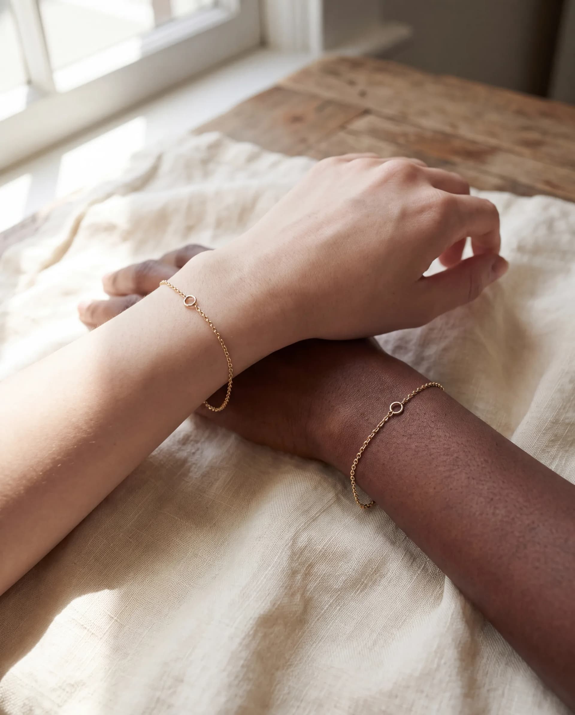 Two wrists resting on ivory linen, each wearing a fine 14K yellow gold chain bracelet closed with a tiny jump ring