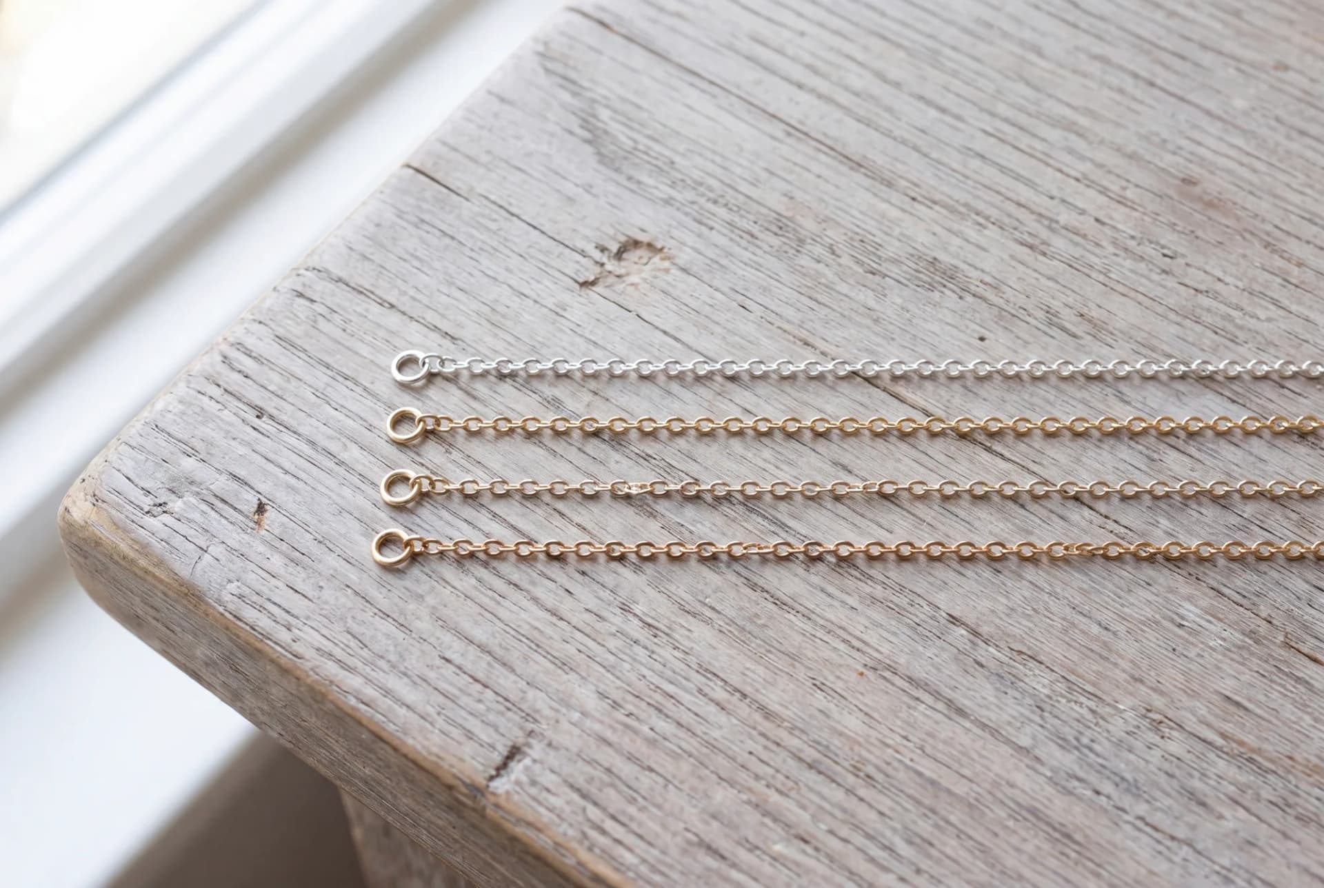 Four fine chains laid flat in a row on pale oak, from sterling silver to 14K yellow gold, each a slightly different tone