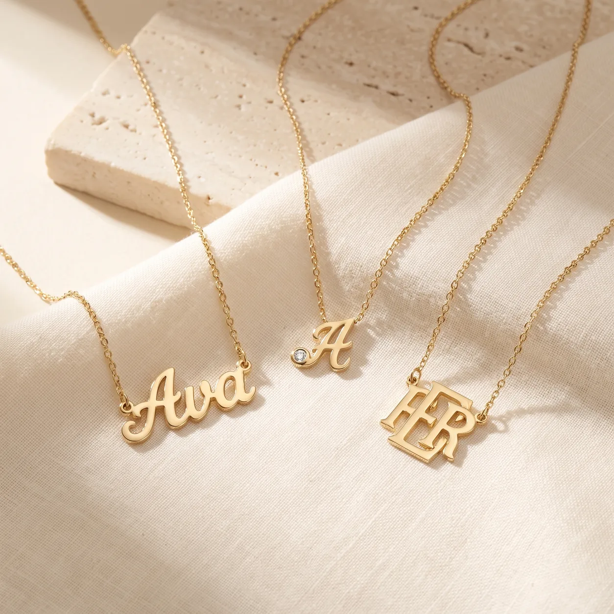 A gold script nameplate necklace reading Ava, a gold script initial A pendant with a diamond accent, and an interlocked two-letter gold monogram pendant on cream linen