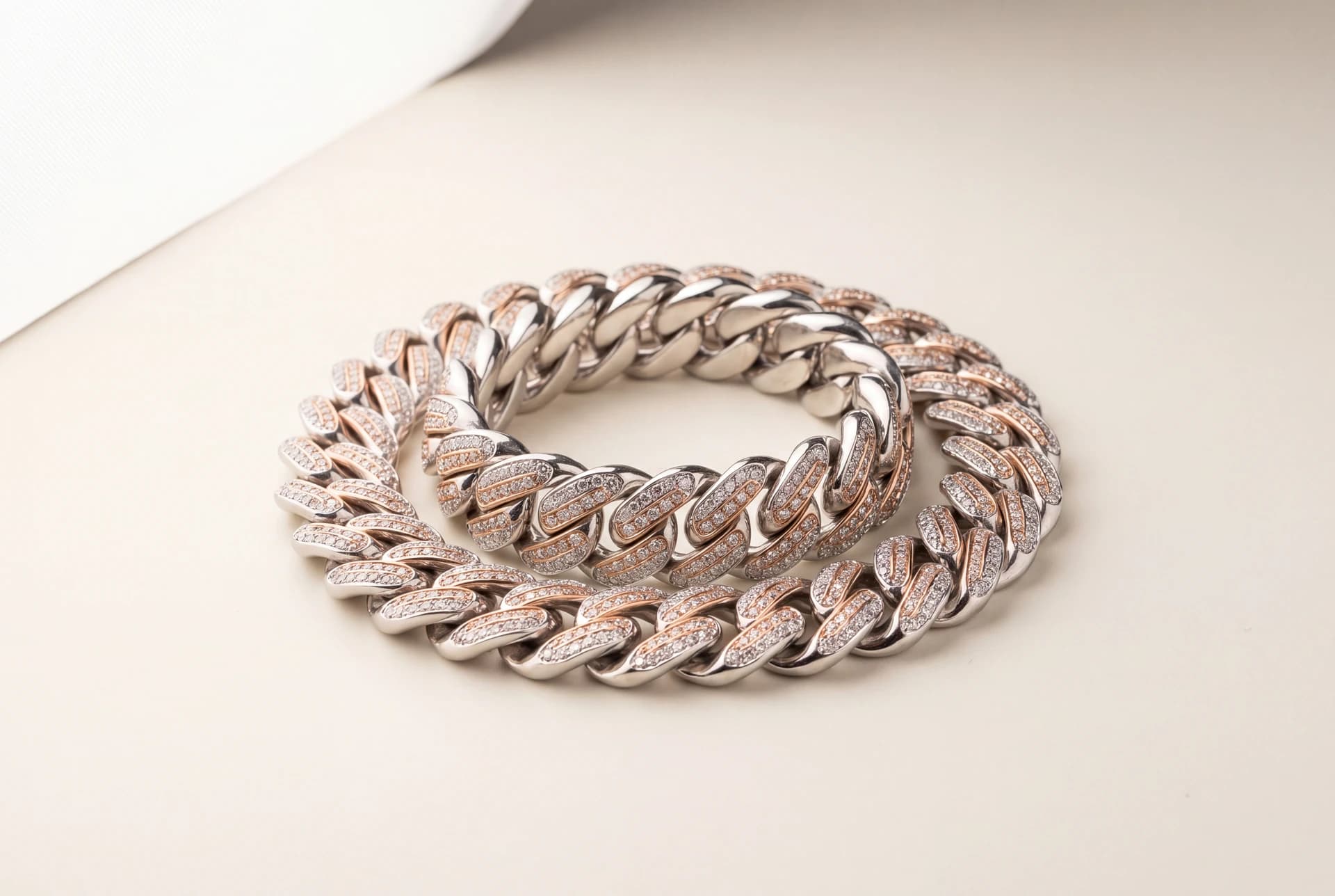 Two coiled iced-out Cuban link chains, one white gold and one rose gold, on a cream surface.