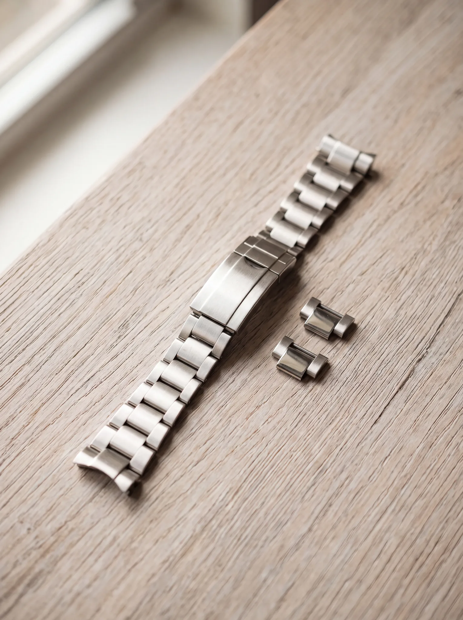 A steel watch bracelet, unclasped, with two loose links beside it, resting on a pale weathered oak table in soft window light at ATL Luxury Jewelry, Snellville GA