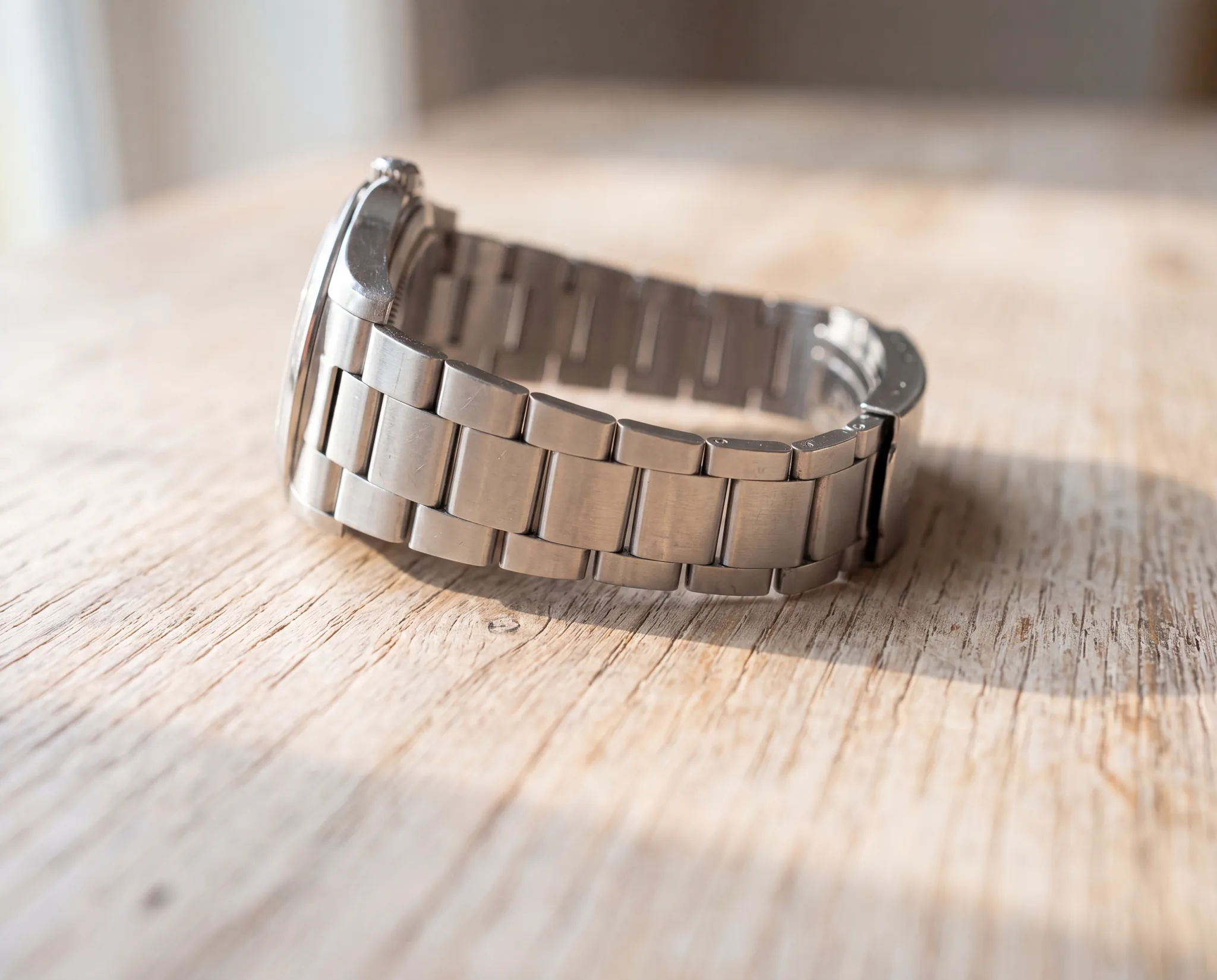 A steel watch bracelet curved into a wide open loop with visible gaps between the links, resting on a pale weathered oak table at ATL Luxury Jewelry, Snellville GA