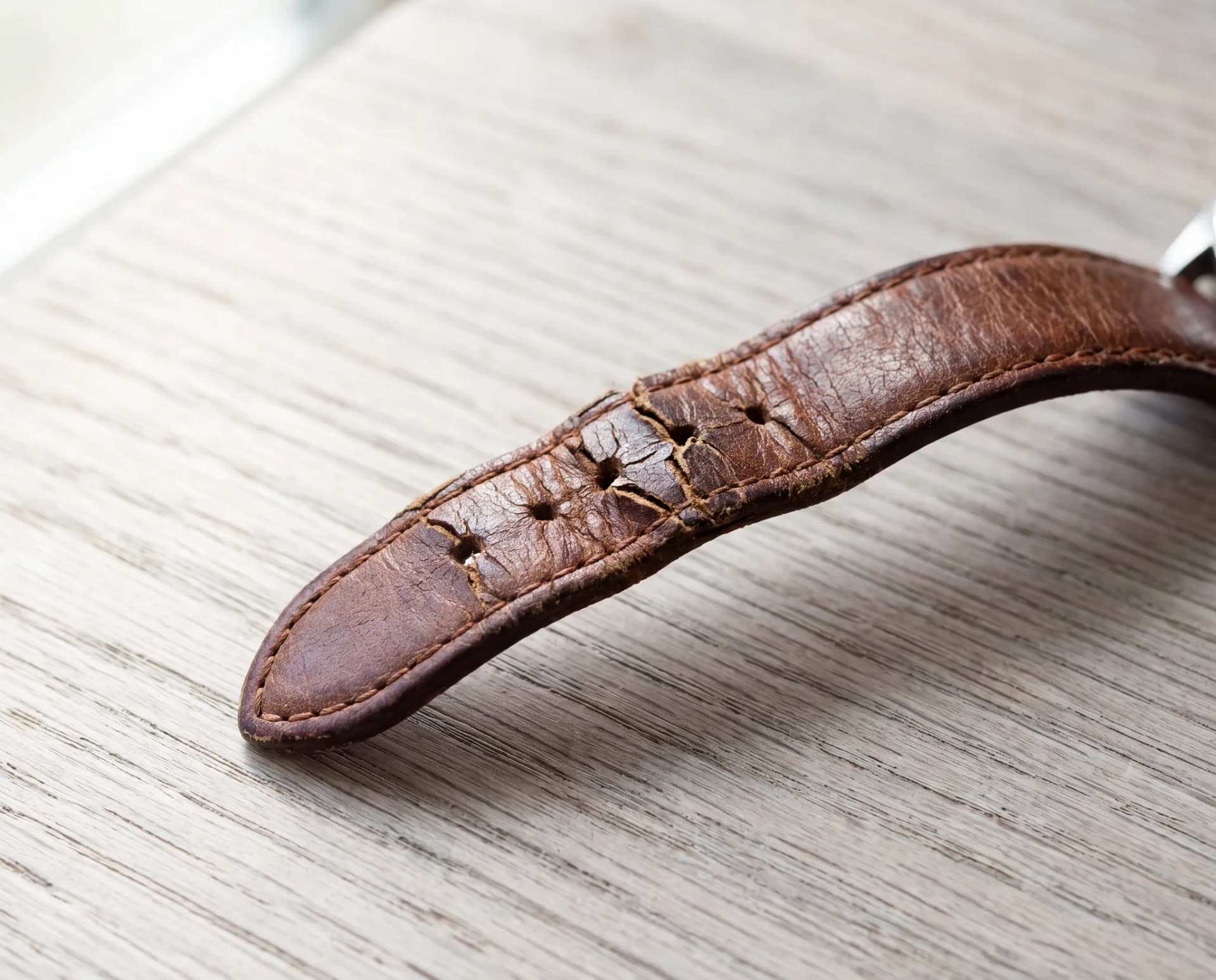 A worn brown leather watch strap with cracked and split leather at the buckle holes, resting on a pale weathered oak table at ATL Luxury Jewelry, Snellville GA