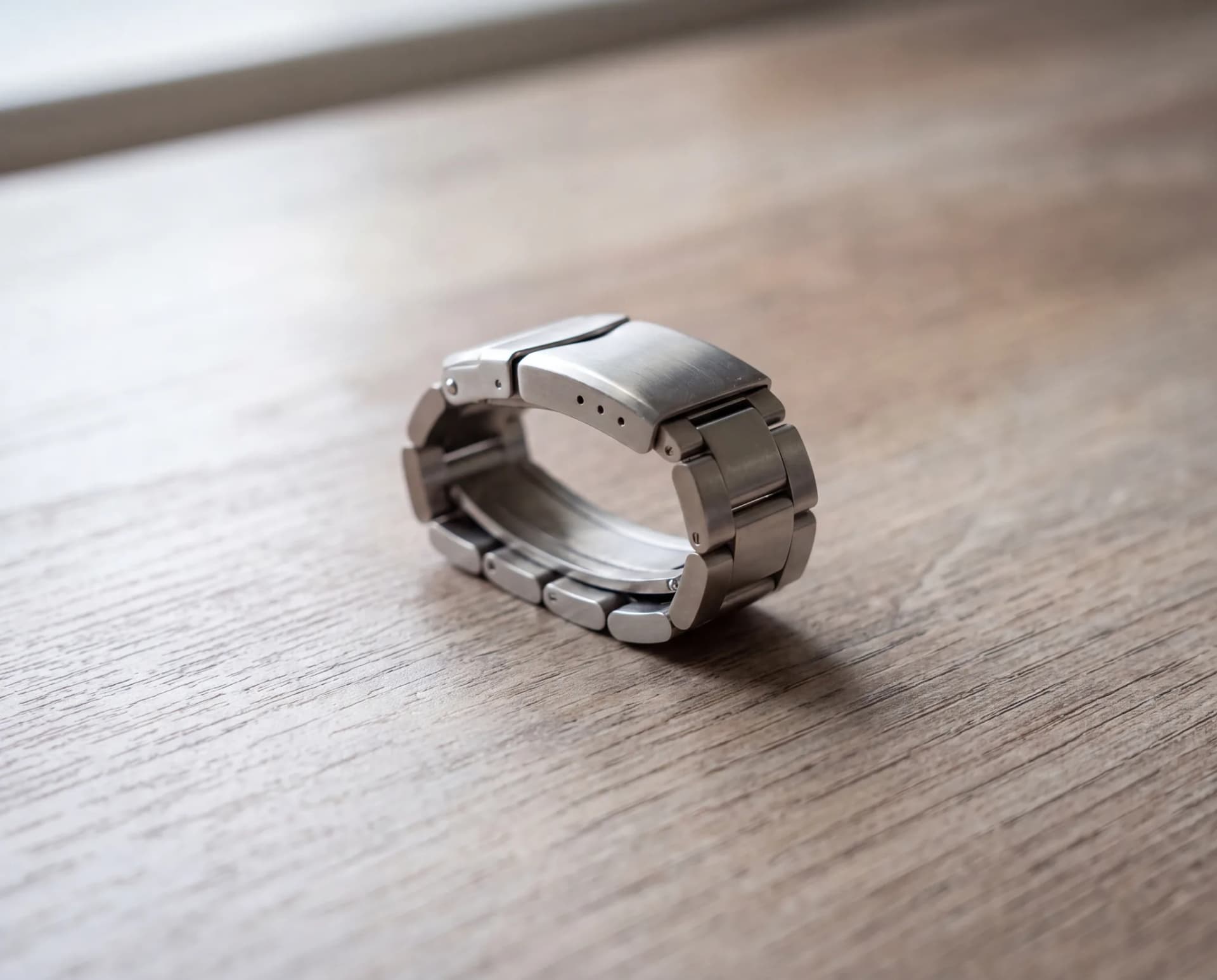 A steel watch bracelet curved into a tight closed loop with its links pressed together, resting on a pale weathered oak table at ATL Luxury Jewelry, Snellville GA