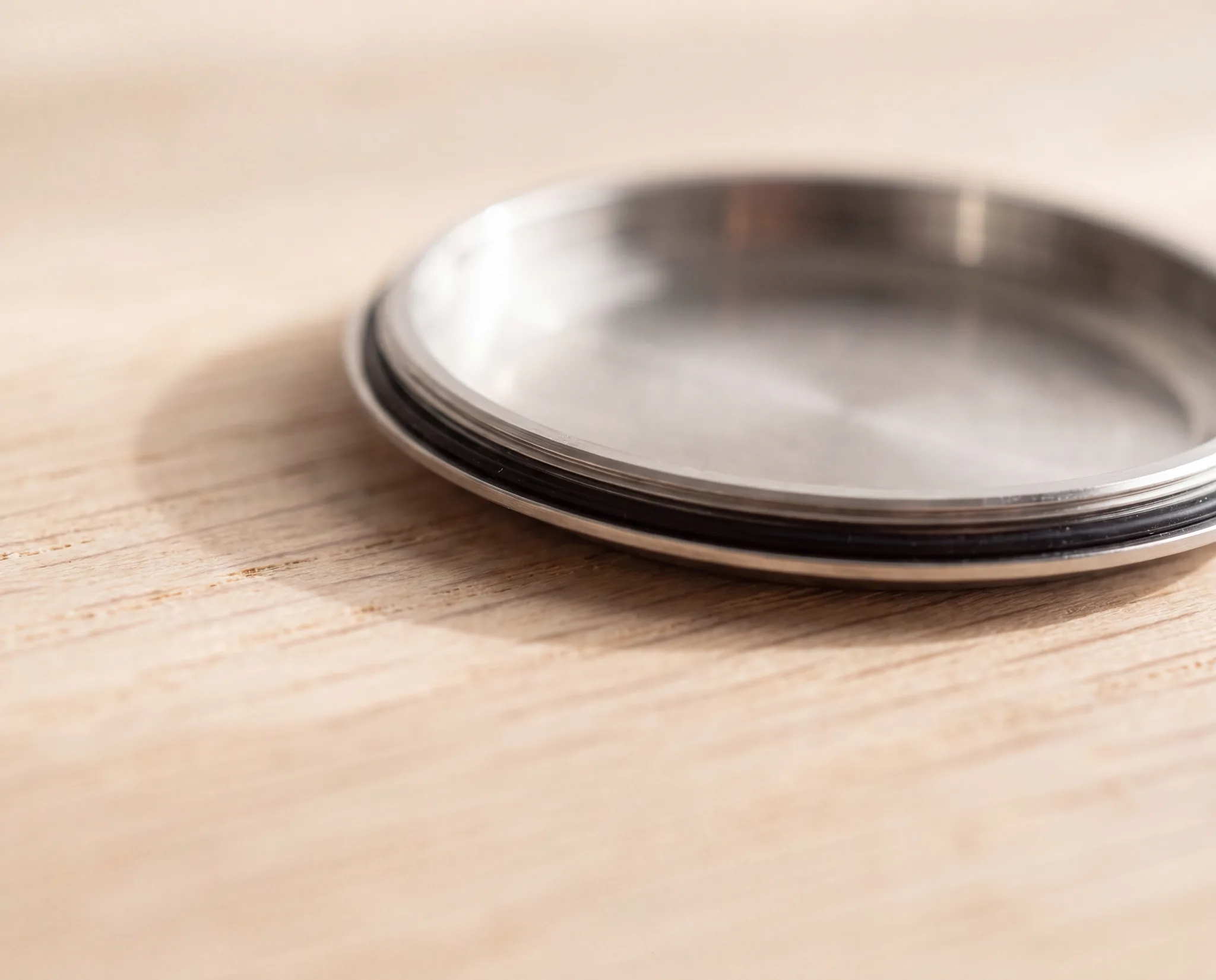 A close-up of a steel watch case back with its black rubber gasket visible around the rim, resting on a pale weathered oak table at ATL Luxury Jewelry, Snellville GA