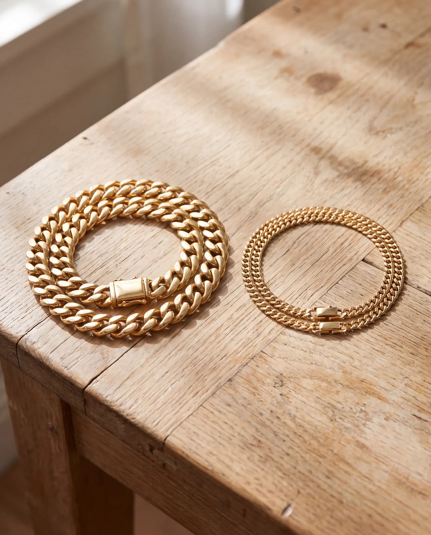 Two solid gold Cuban link chains coiled on a pale oak wood surface