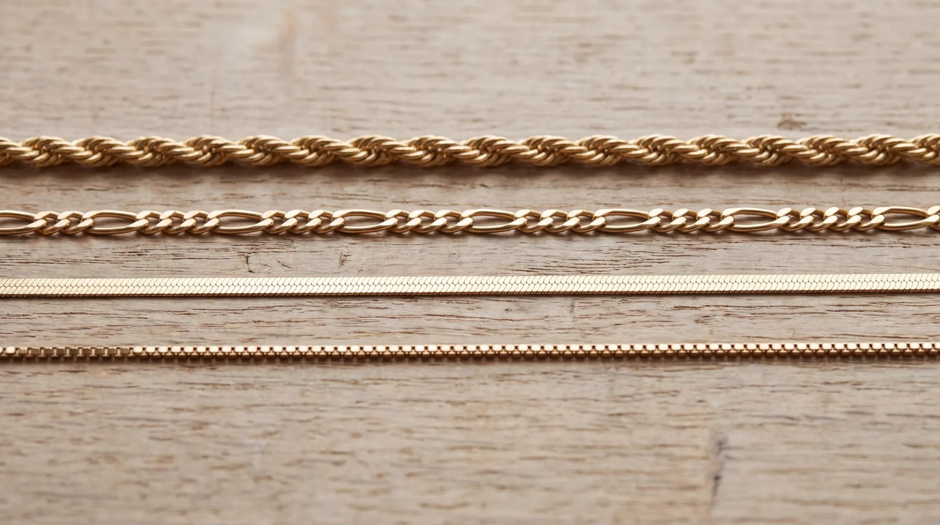 Rope, figaro, herringbone and box chains laid out together on a pale oak wood surface