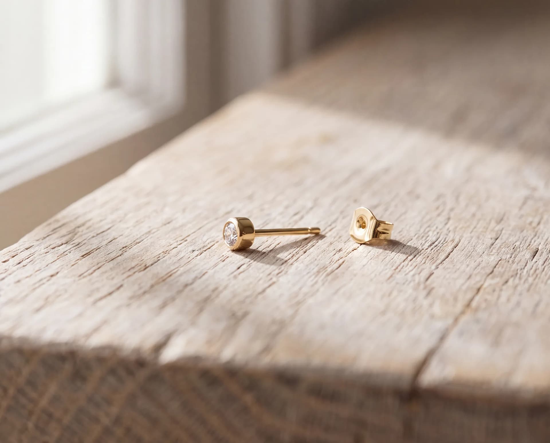 A gold stud earring on a pale wood surface with its butterfly back lying loose a short distance away