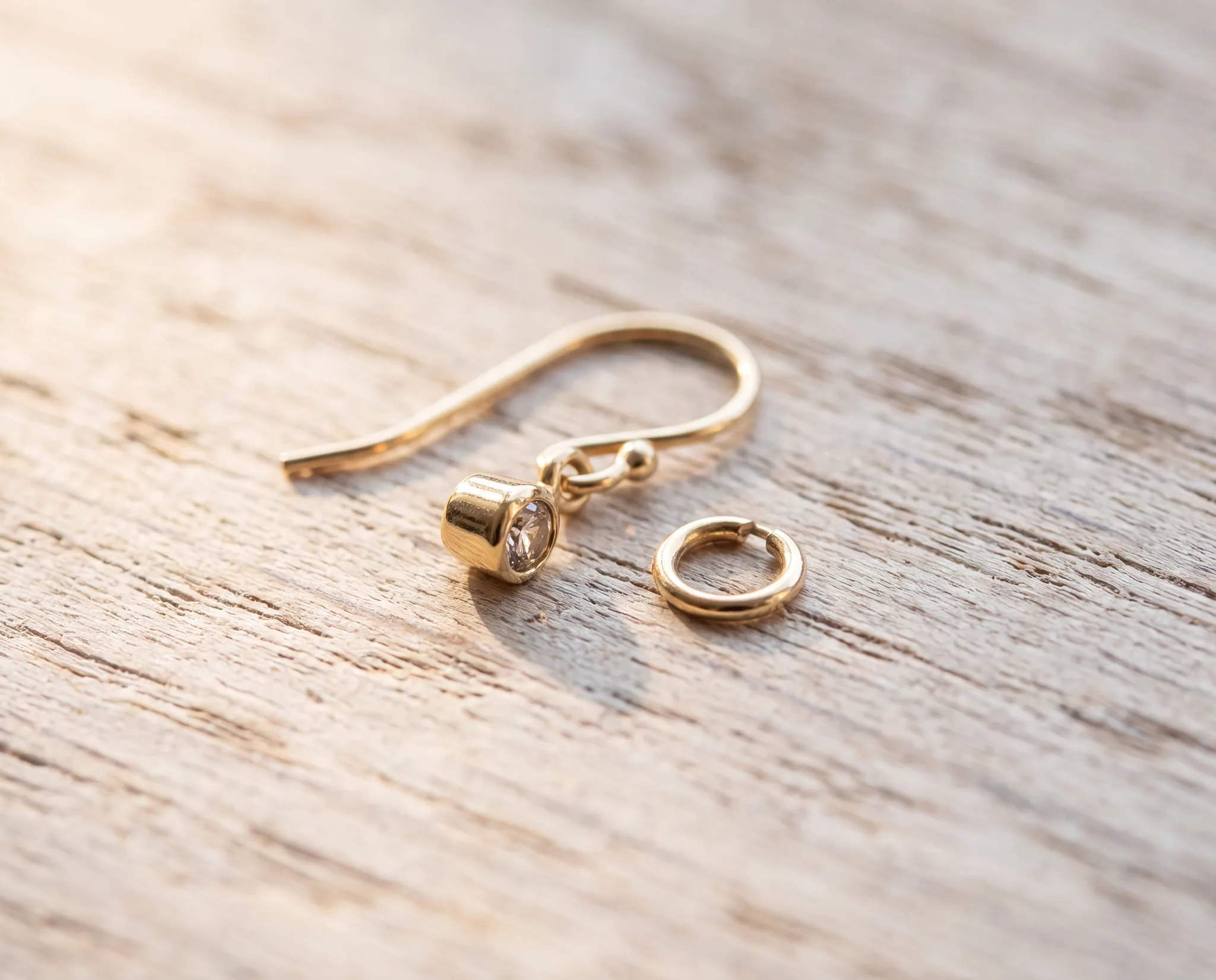 A gold drop earring on a pale wood surface with its dangle detached beside the open jump ring it came off