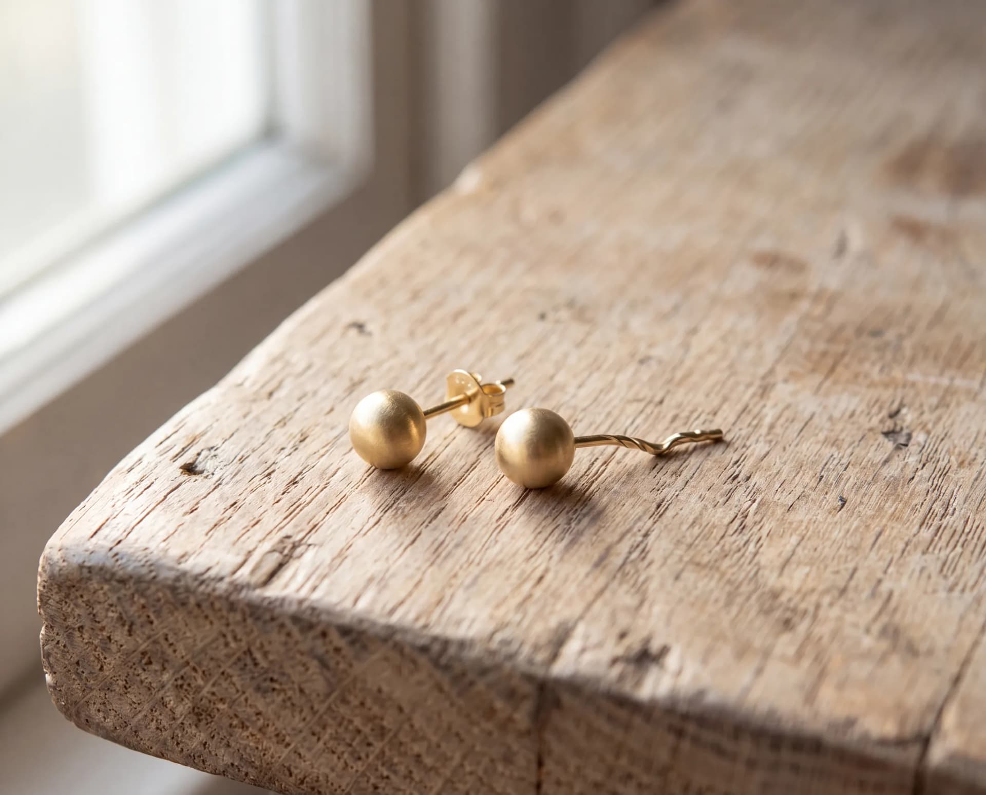 Two matching gold stud earrings on a pale wood surface, one undamaged and one with a bent post