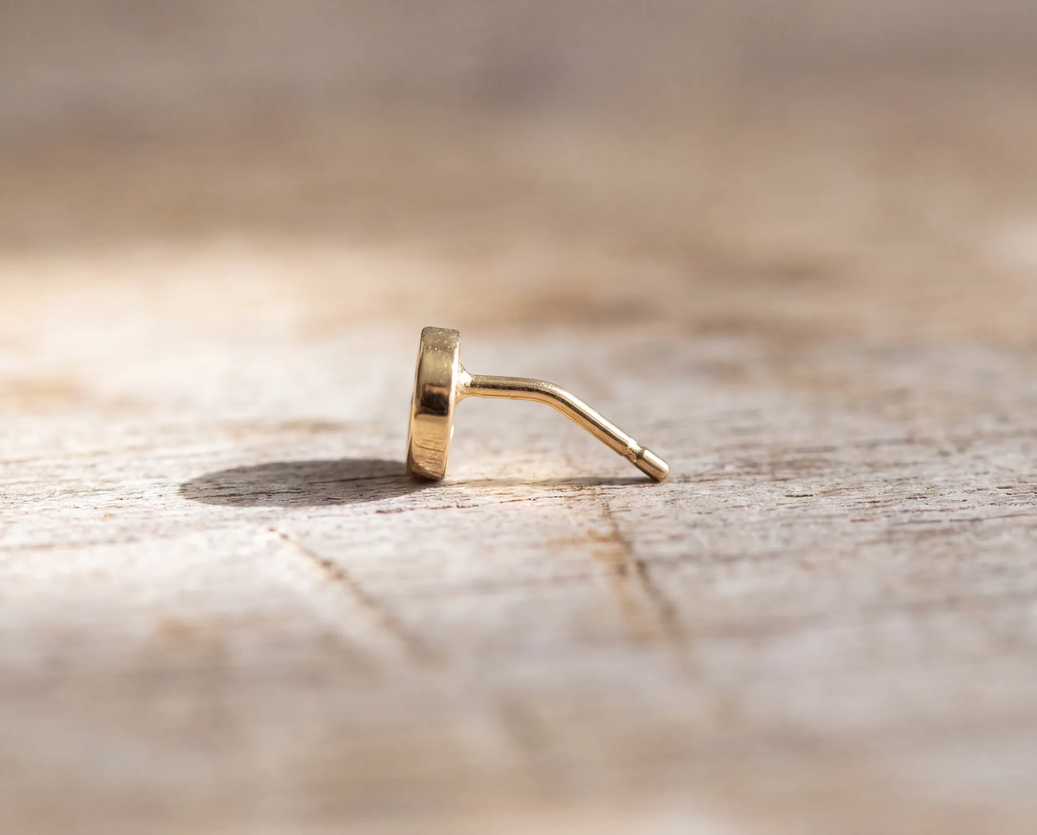 A gold stud earring lying on its side on a pale wood surface with its post bent at an angle