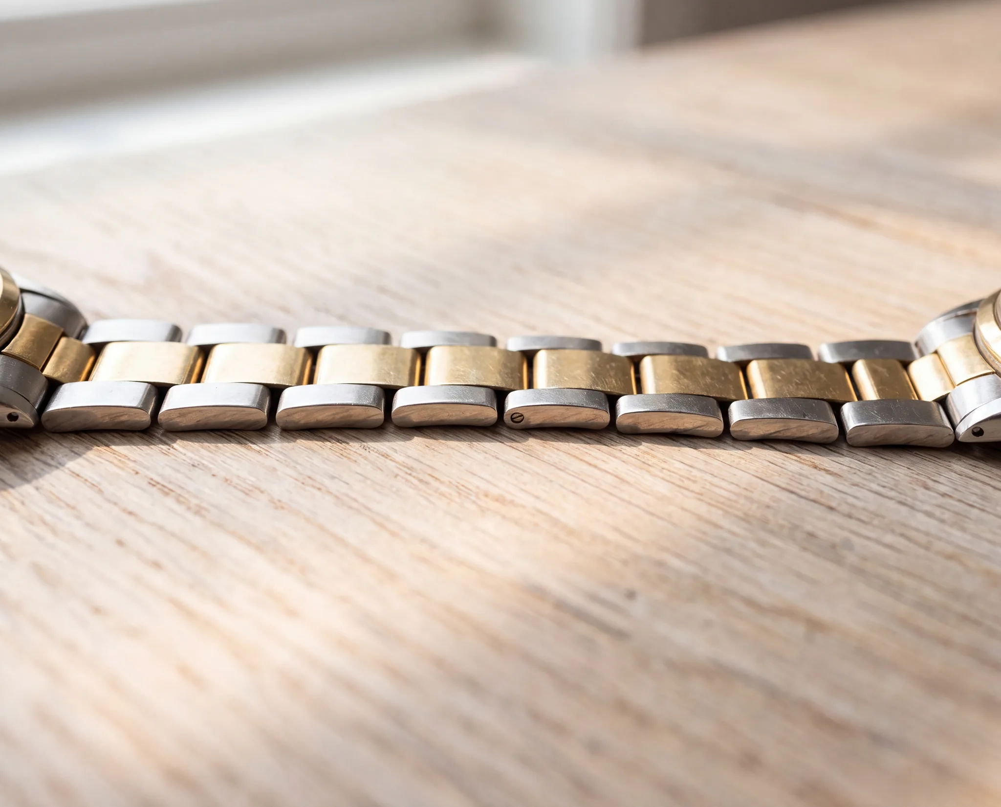 A two-tone steel and gold watch bracelet lying open and extended on a pale weathered oak table, its individual links visible