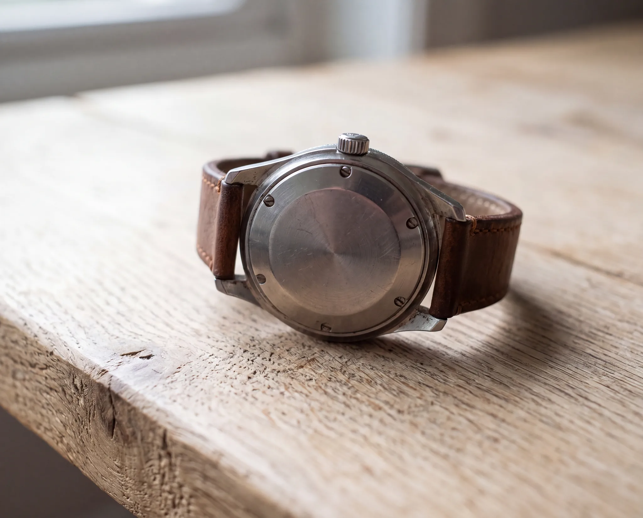 The steel case back of a wristwatch on a brown leather strap, shown from behind with its screws visible, resting on a pale weathered oak table near a window