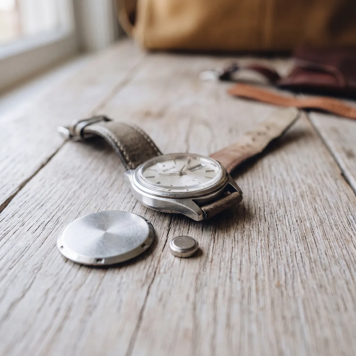 A wristwatch with its case back removed, resting beside a small round battery on a pale weathered oak table at ATL Luxury Jewelry, Snellville GA