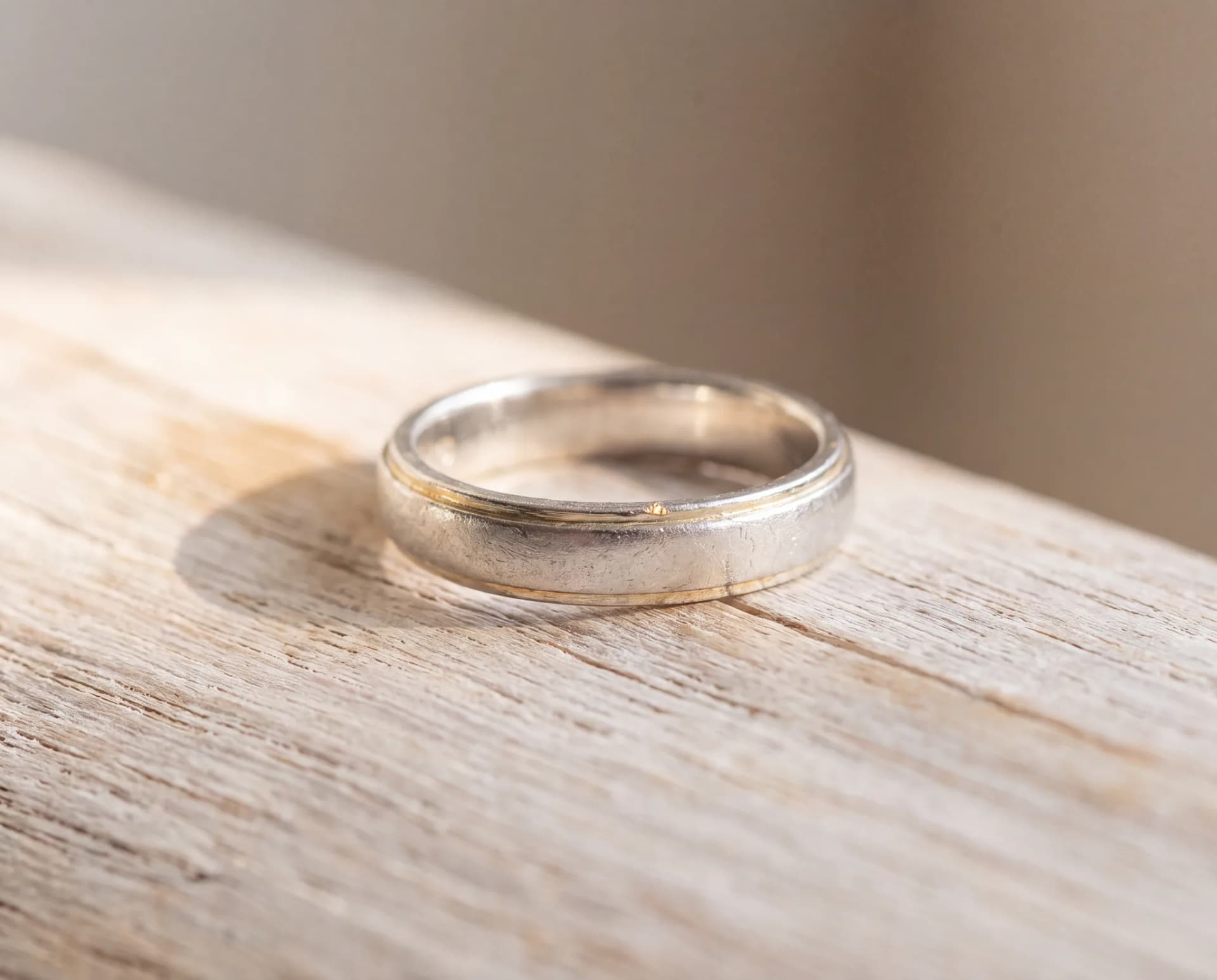 A white gold ring on a pale wood surface that has gone warm and faintly yellow where its plating has worn