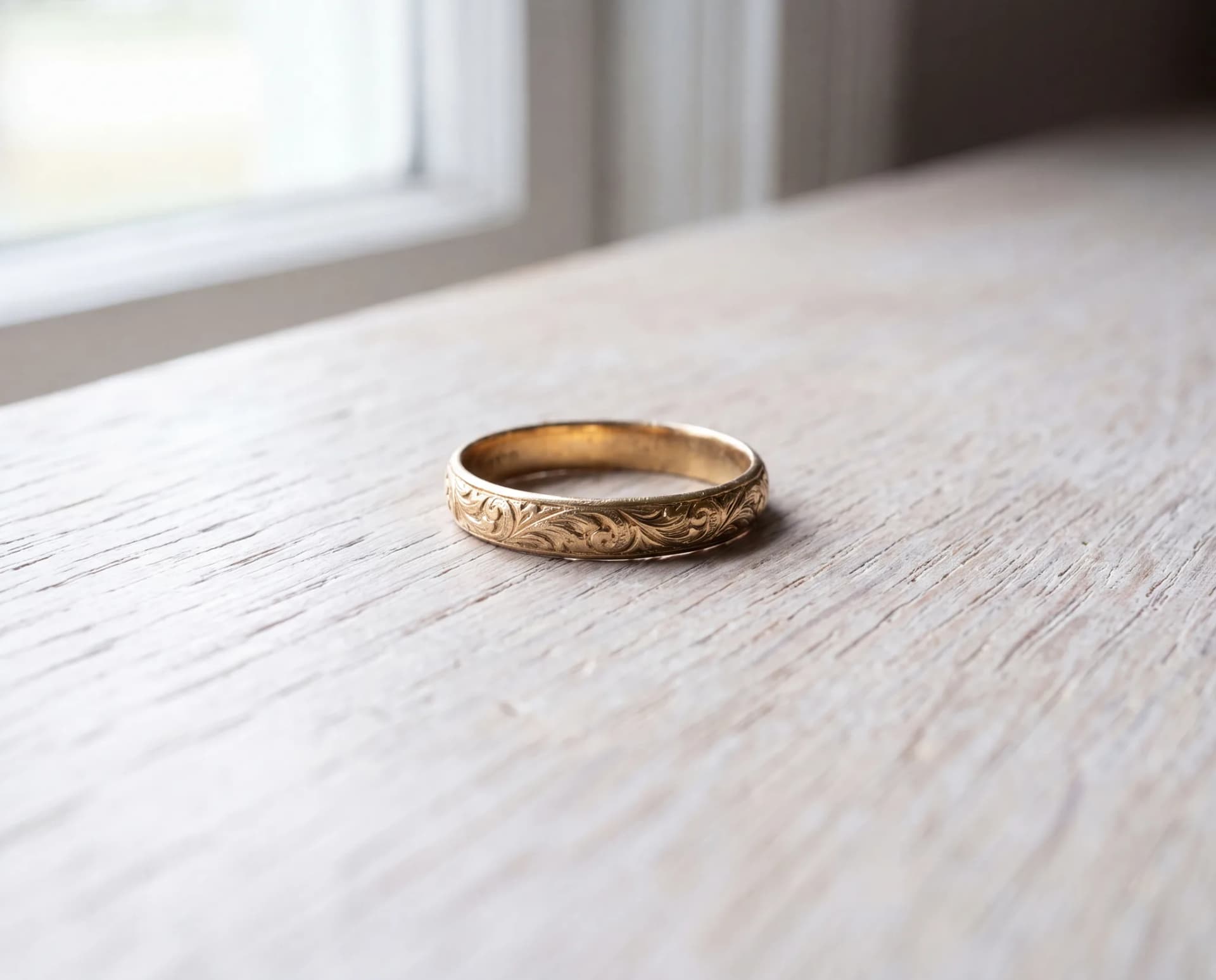An antique engraved gold band with worn filigree detail resting on a pale wood surface