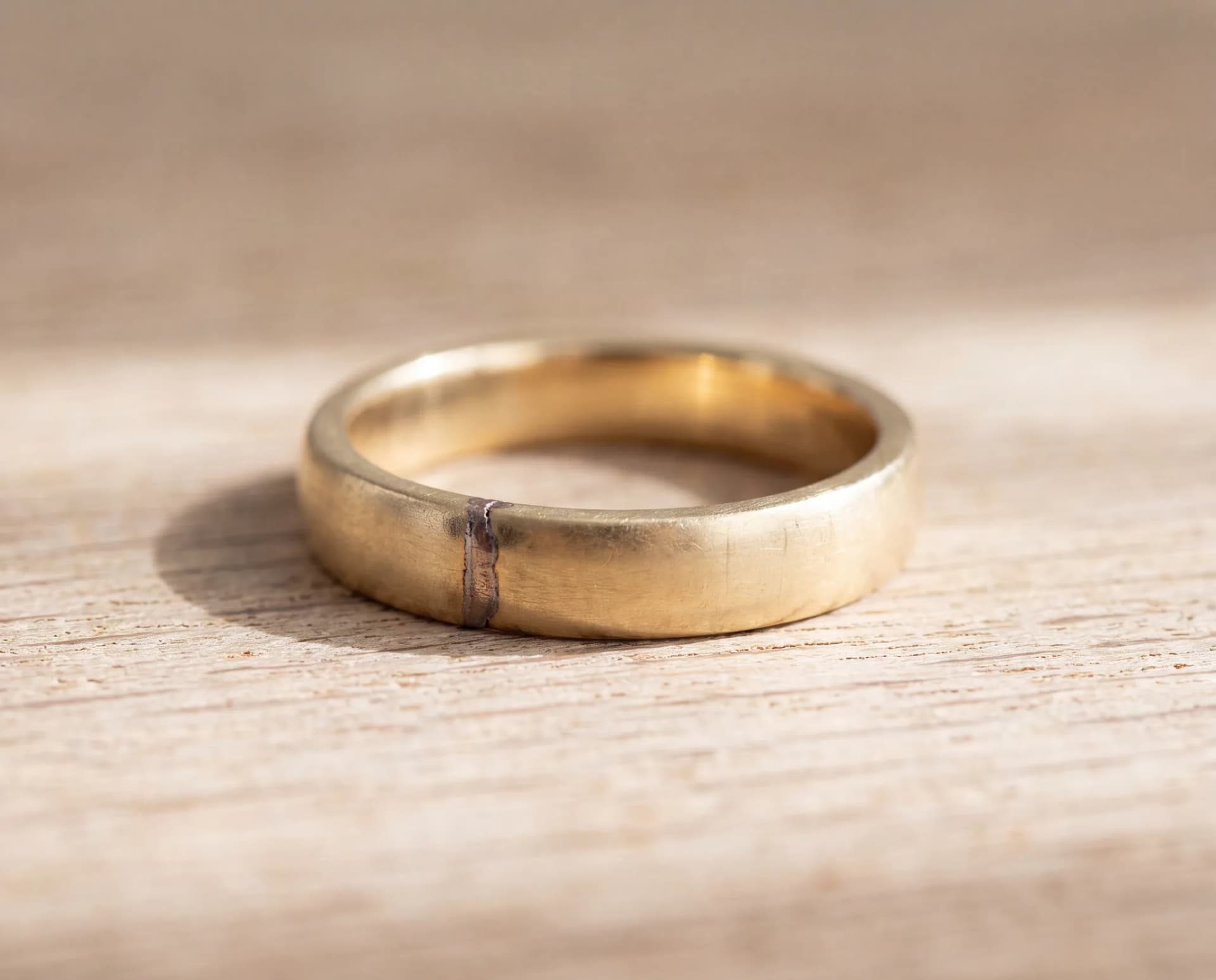 A gold band on a pale wood surface showing a visible solder seam across the shank from an old repair