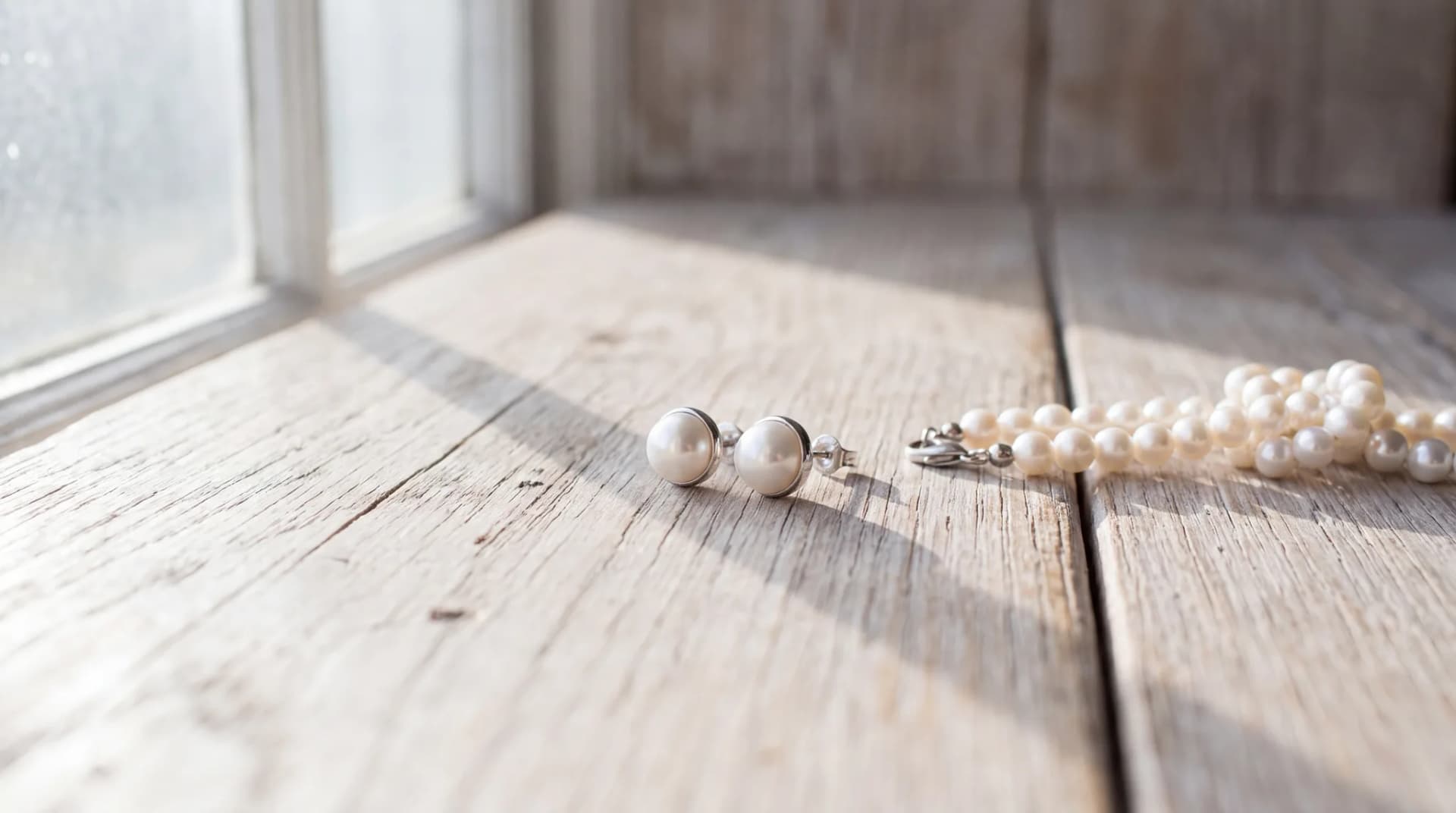 A pair of white pearl stud earrings beside a knotted pearl strand on a pale wood surface
