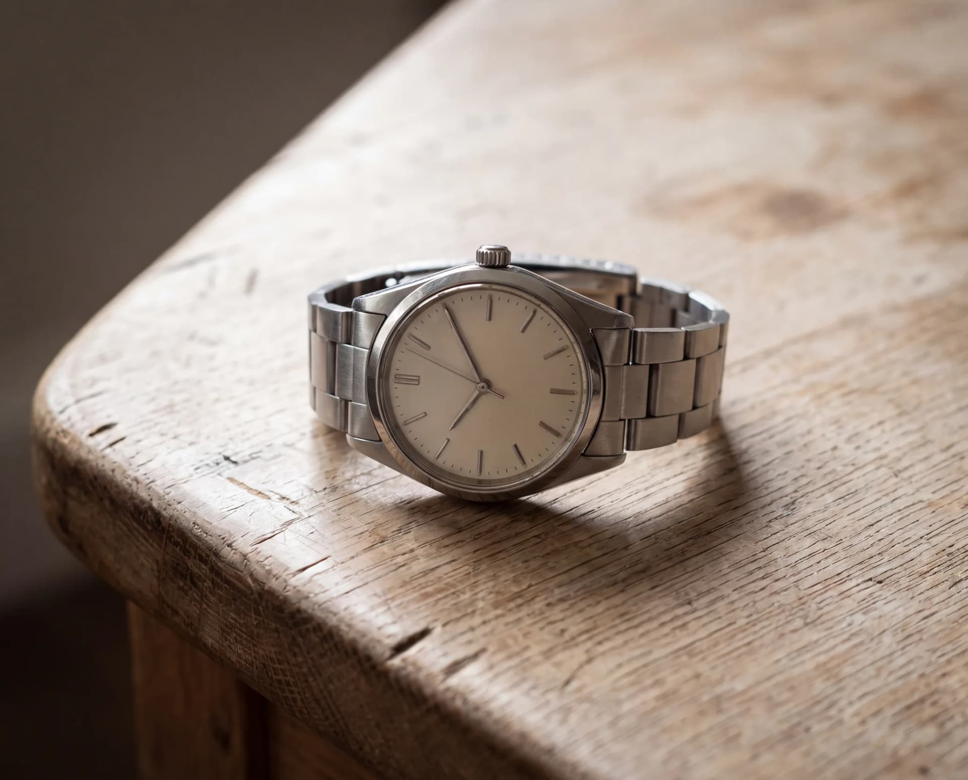 A steel wristwatch with a cream dial, baton hour markers and a matching steel bracelet, lying on a pale weathered oak table at ATL Luxury Jewelry, Snellville GA