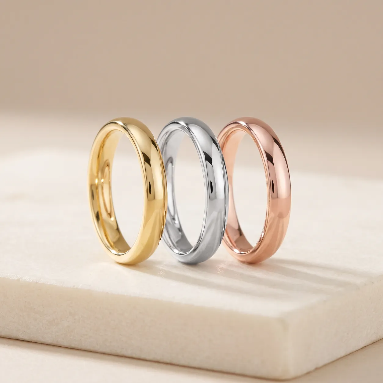 Three plain polished bands standing side by side on a pale stone slab: one yellow gold, one white gold, one rose gold.