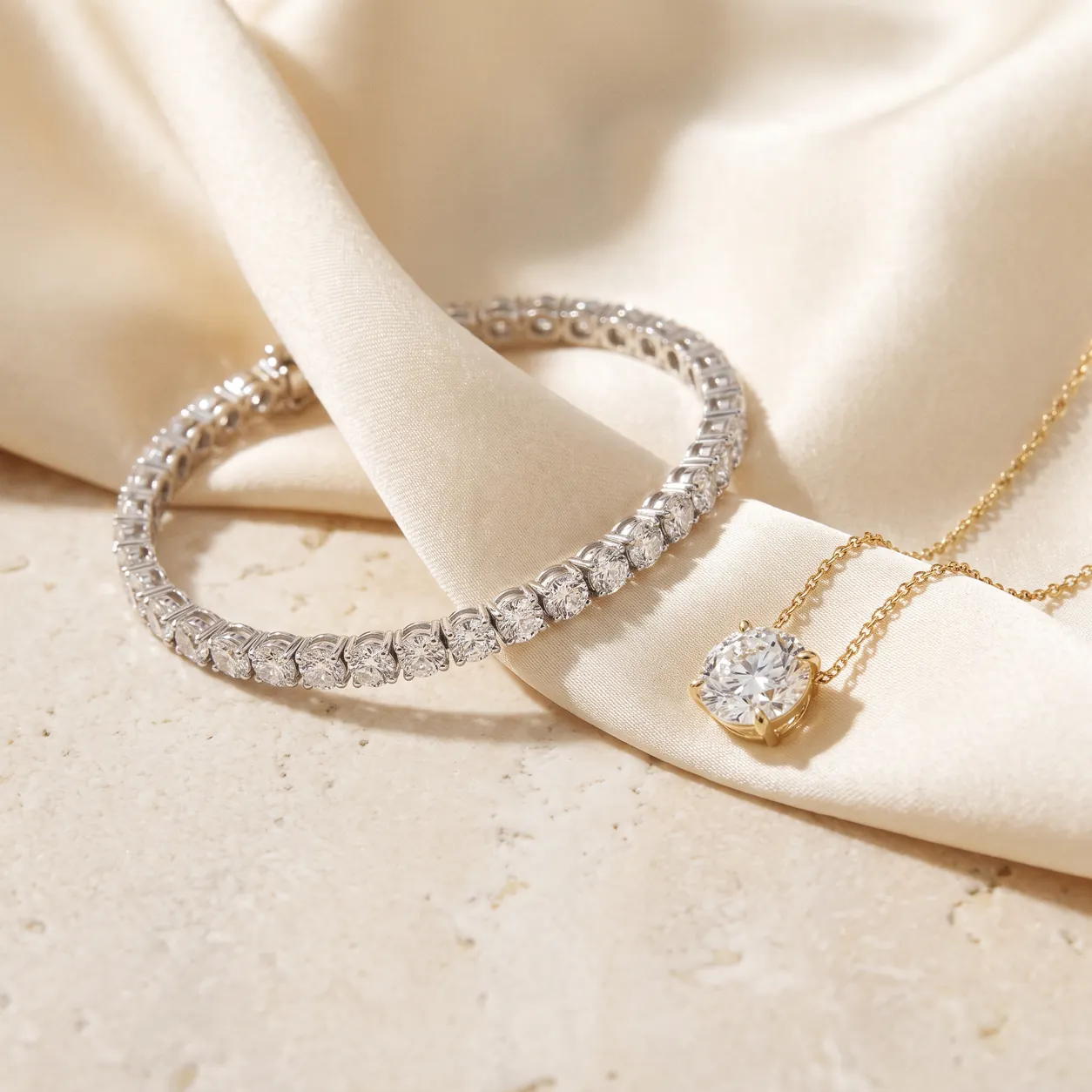 A white-metal diamond tennis bracelet curved on cream silk beside a single round diamond pendant on a fine yellow gold chain.