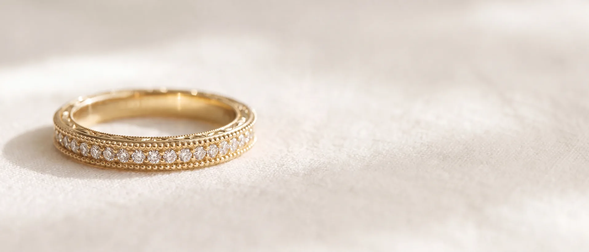 A yellow gold band with beaded milgrain edges, engraved detail on its side and a row of small diamonds, resting on cream fabric